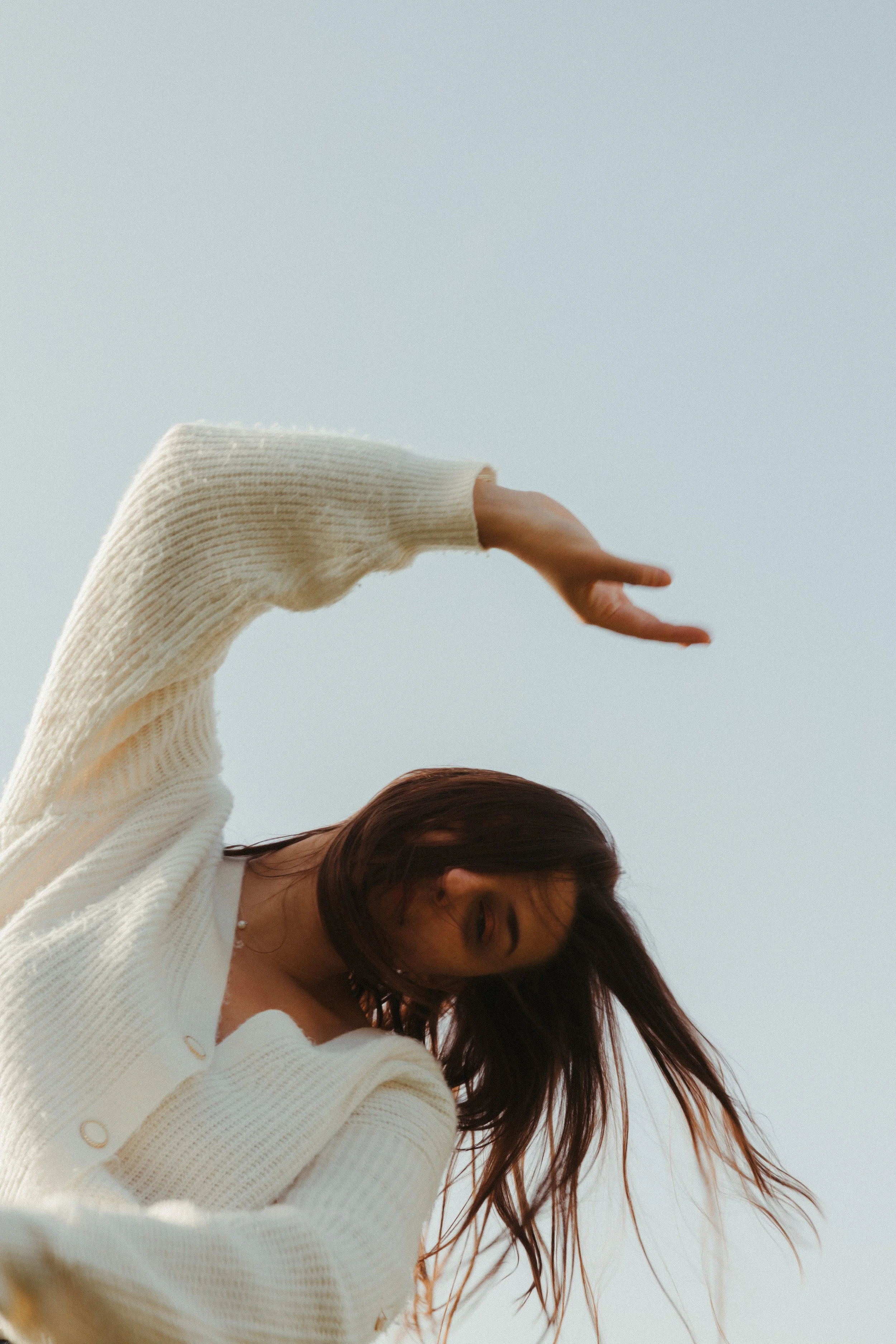 A woman with brown hair, wearing a cream-colored sweater, is looking down while her hair moves in the wind against a light blue sky.