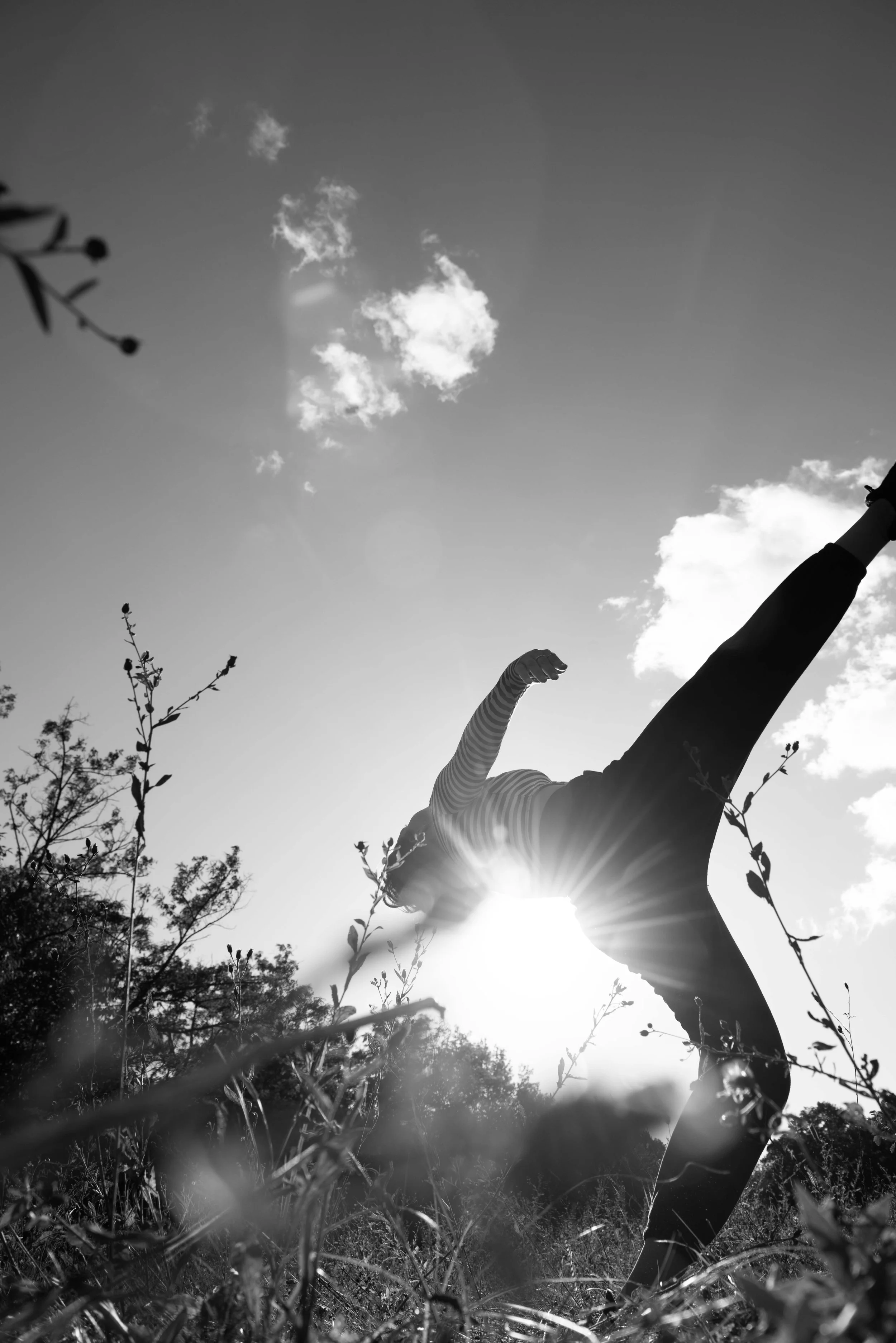 A person outdoors stands in a pose with one arm extended upward and the other arm bent. The sun is shining behind their face, creating a silhouette effect, with a few clouds in the sky and trees in the background.