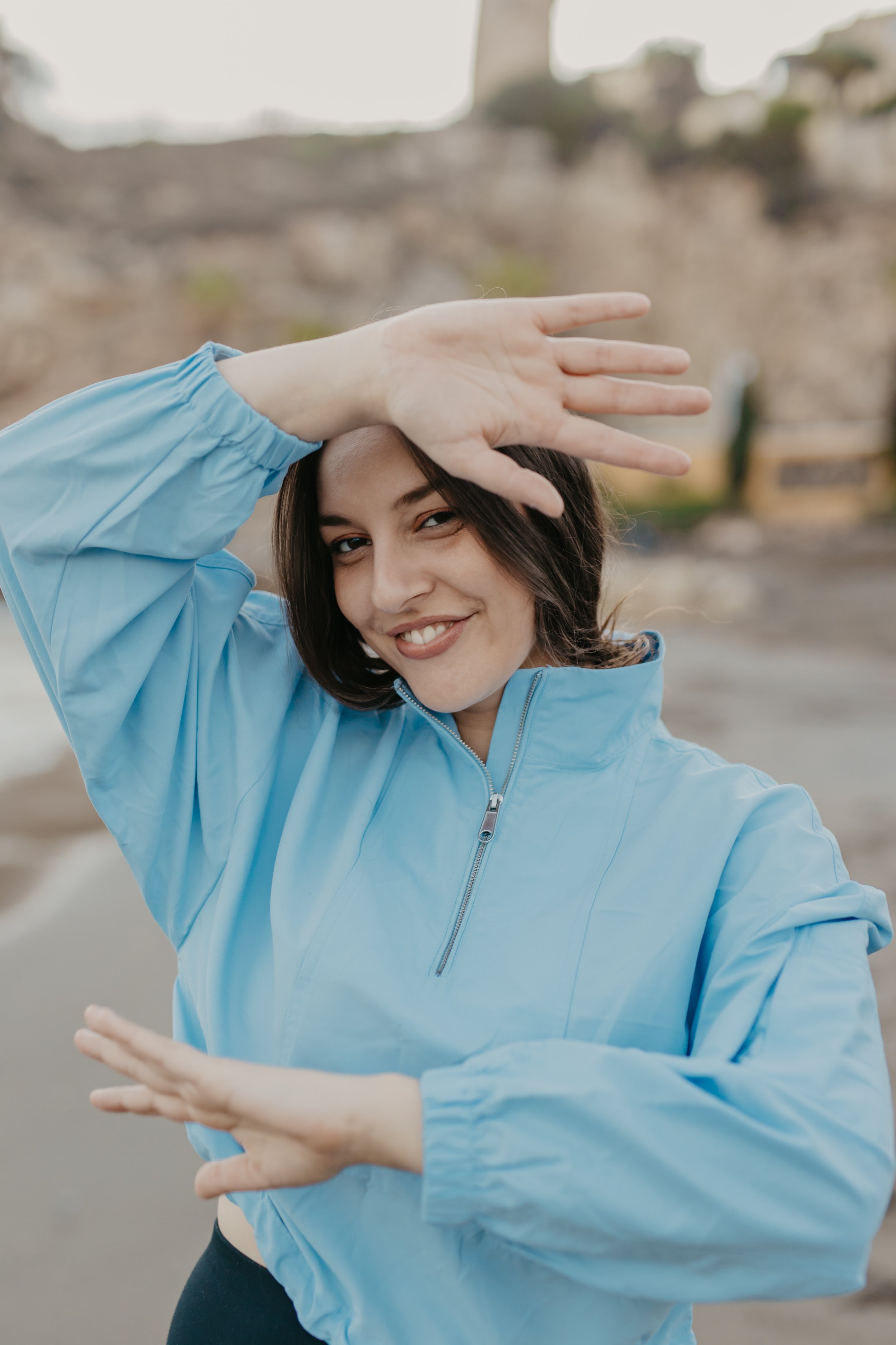 Young woman smiling and posing outdoors in a light blue windbreaker, covering one eye with her hand.