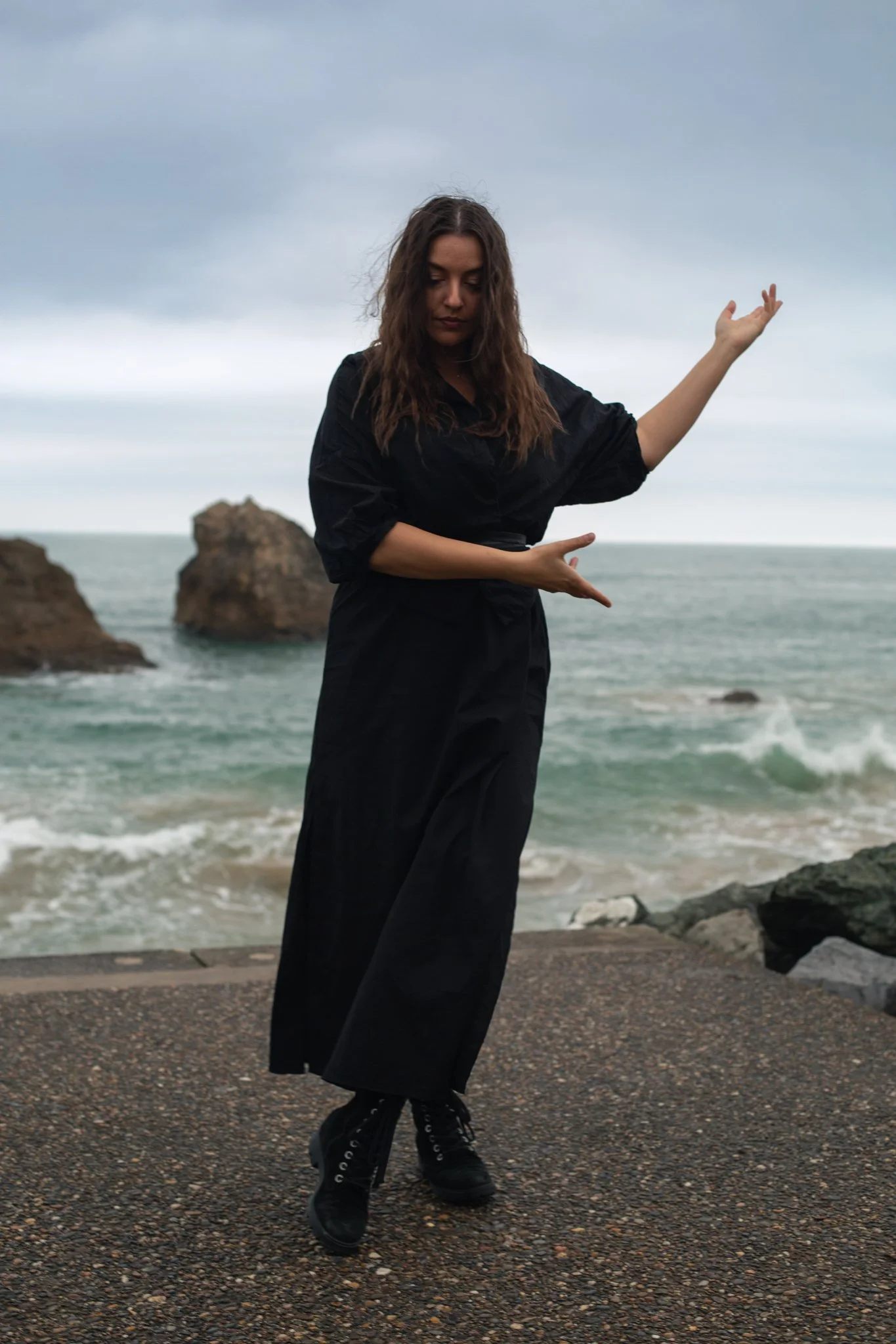 A woman in black standing on a rocky shoreline with waves and rock formations in the background, under a cloudy sky.