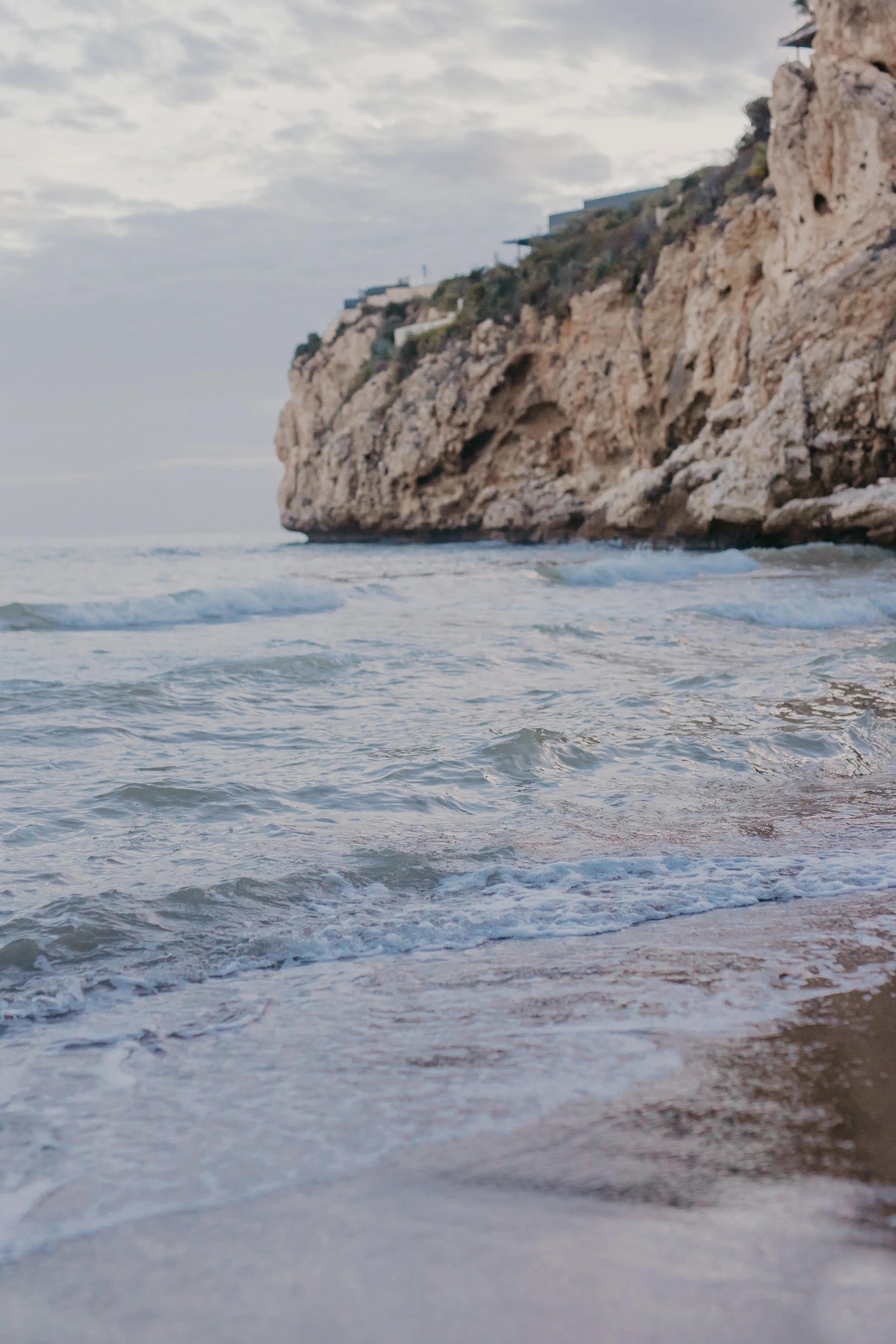 Photo of a rocky seaside cliff with waves crashing on the sandy beach below, under a cloudy sky.