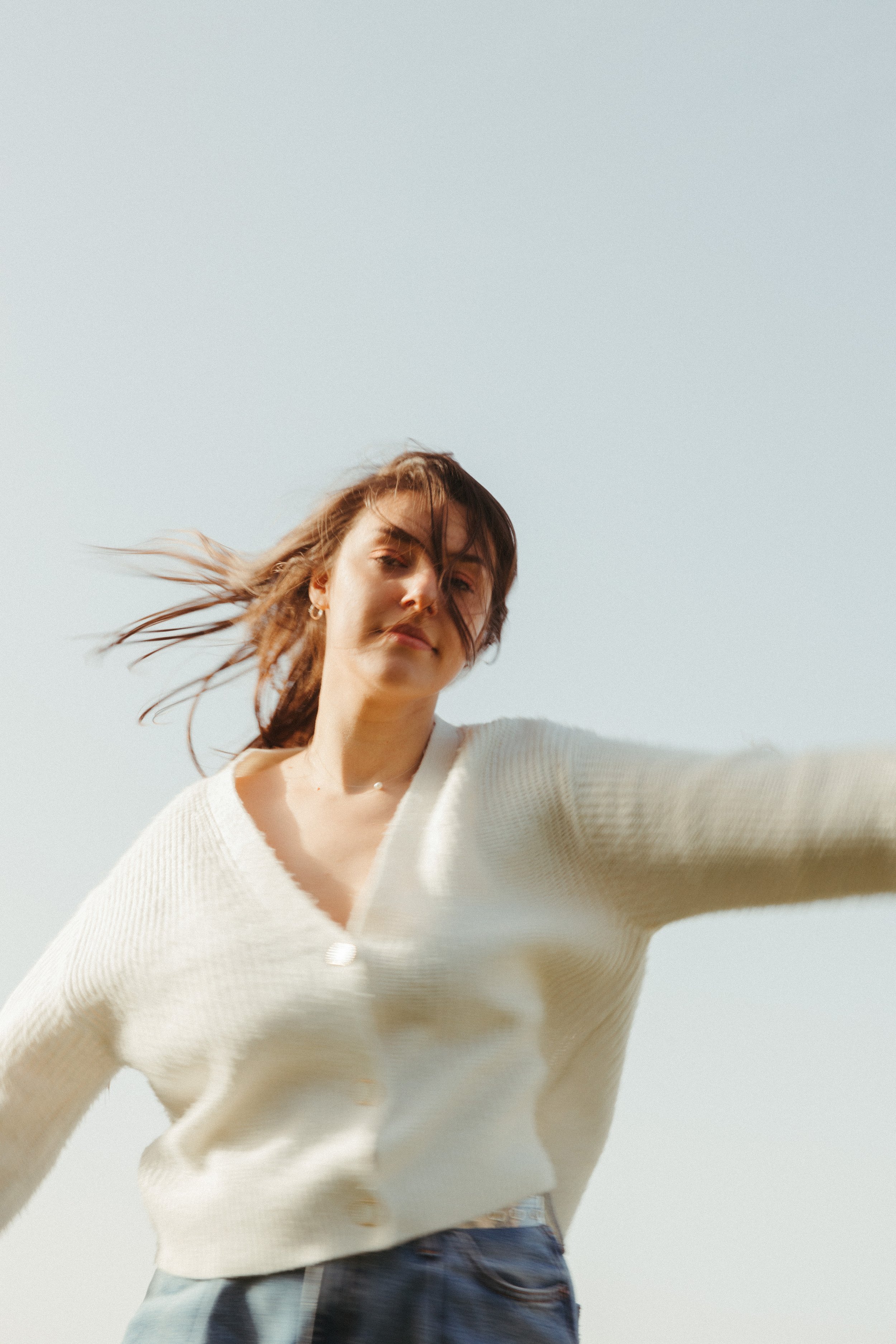 A woman with brown hair in a white sweater and blue jeans standing outdoors against a clear sky, with her hair blowing in the wind.