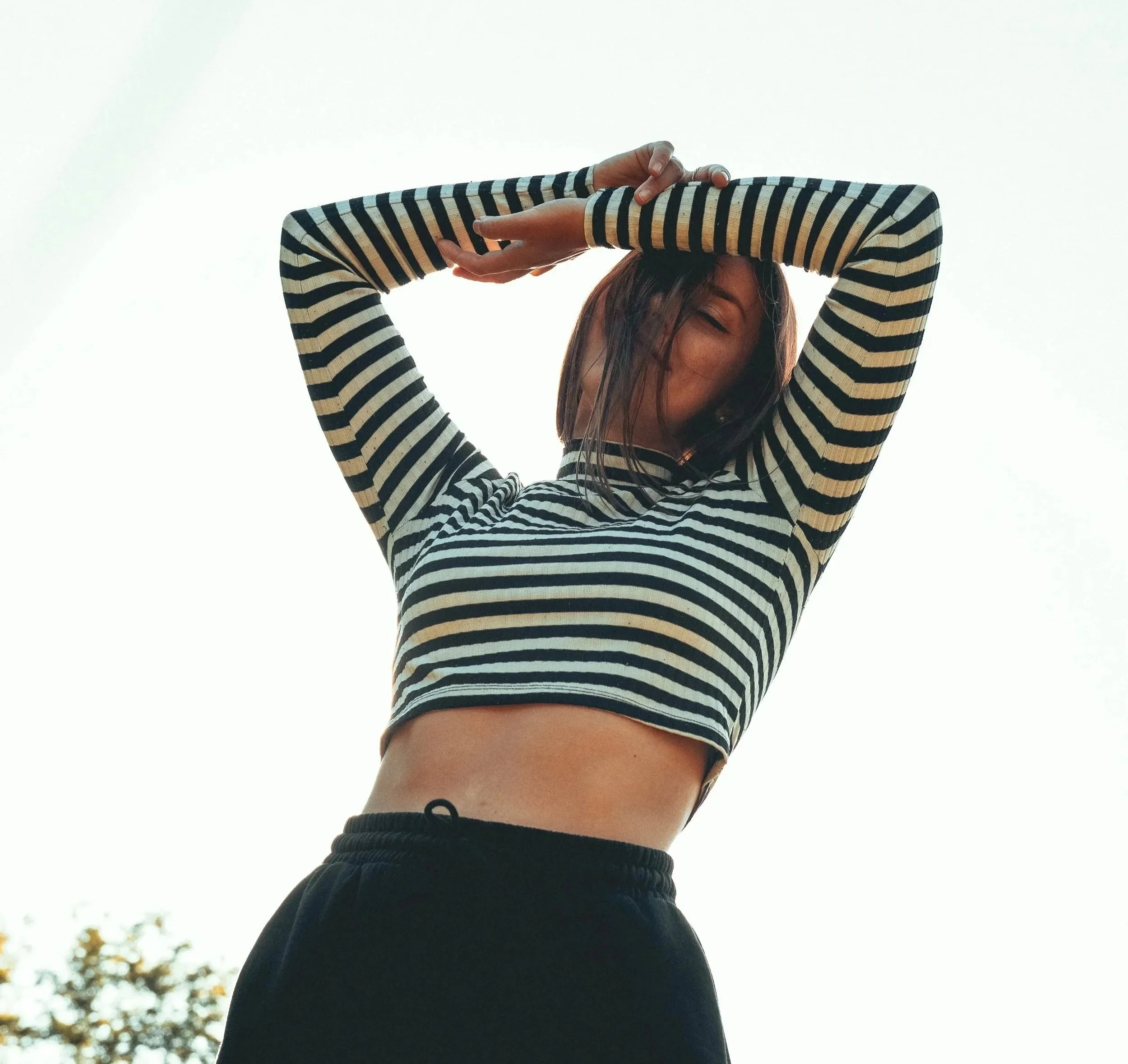 A woman with short hair wearing a black and white striped long-sleeve crop top and black pants, standing outdoors against a bright sky, with her arms raised above her head.