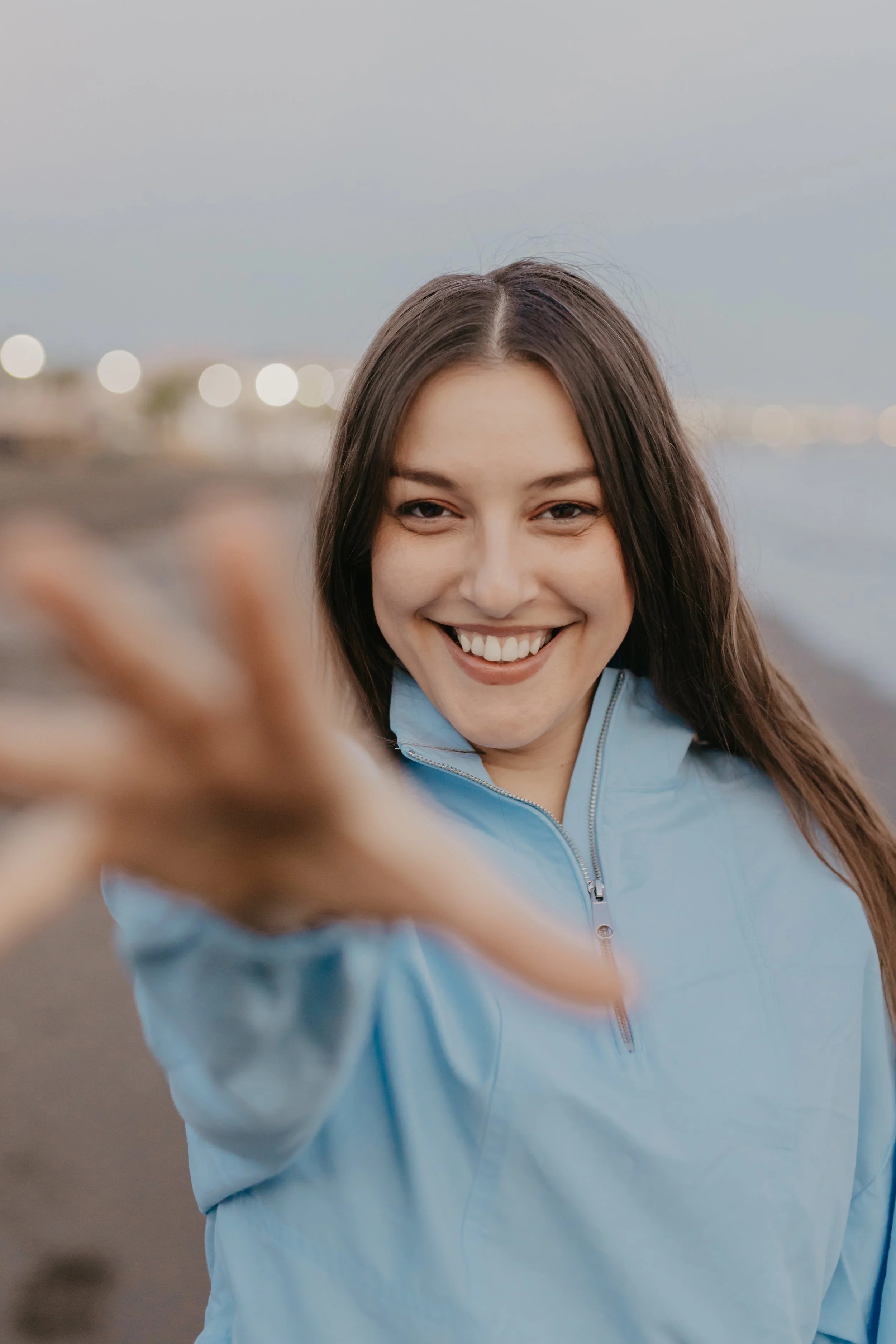 A woman smiling and reaching out her hand towards the camera on a beach at dusk.