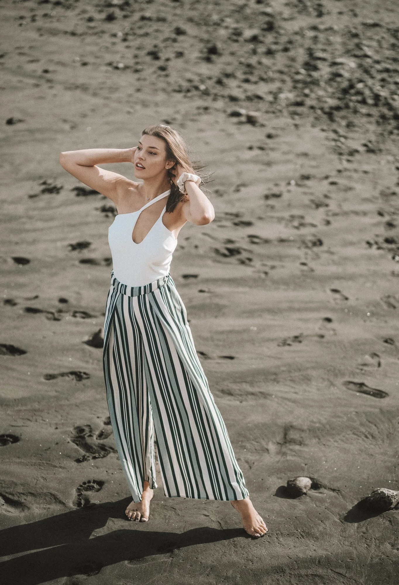 A young woman in striped wide-leg pants and a white top standing barefoot on sandy beach with rocks, posing with her hands in her hair.
