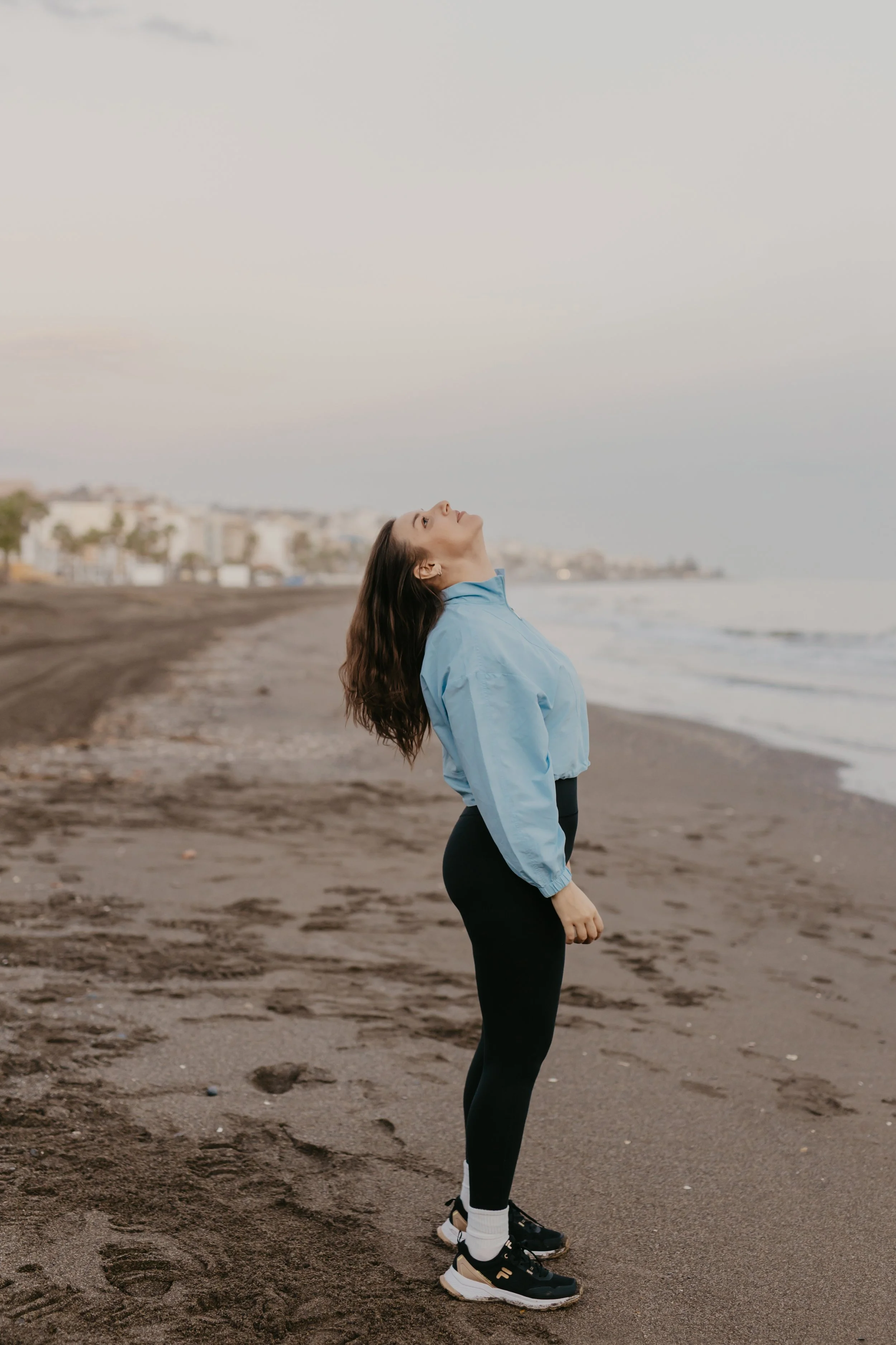 A woman standing on a sandy beach near the ocean, wearing a blue jacket, black leggings, white socks, and black sneakers, looking upwards with her eyes closed.