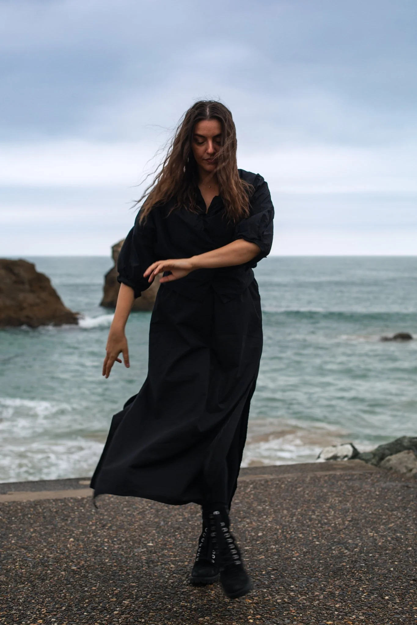 A woman dressed in black standing on a rocky seashore with ocean and cloudy sky in the background.