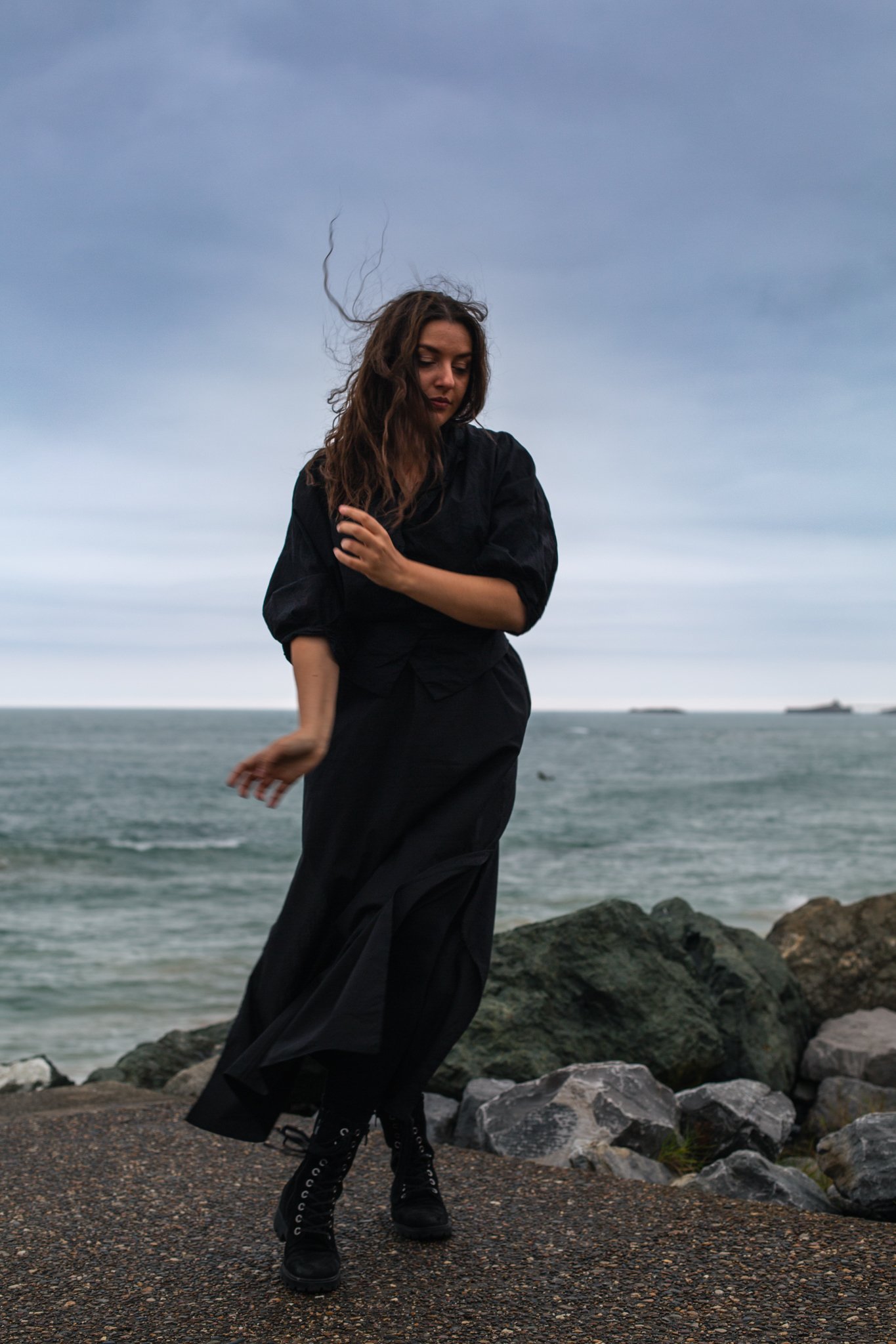 A woman standing on rocky terrain near water, wearing a black outfit, with wind-blown hair, under a cloudy sky.