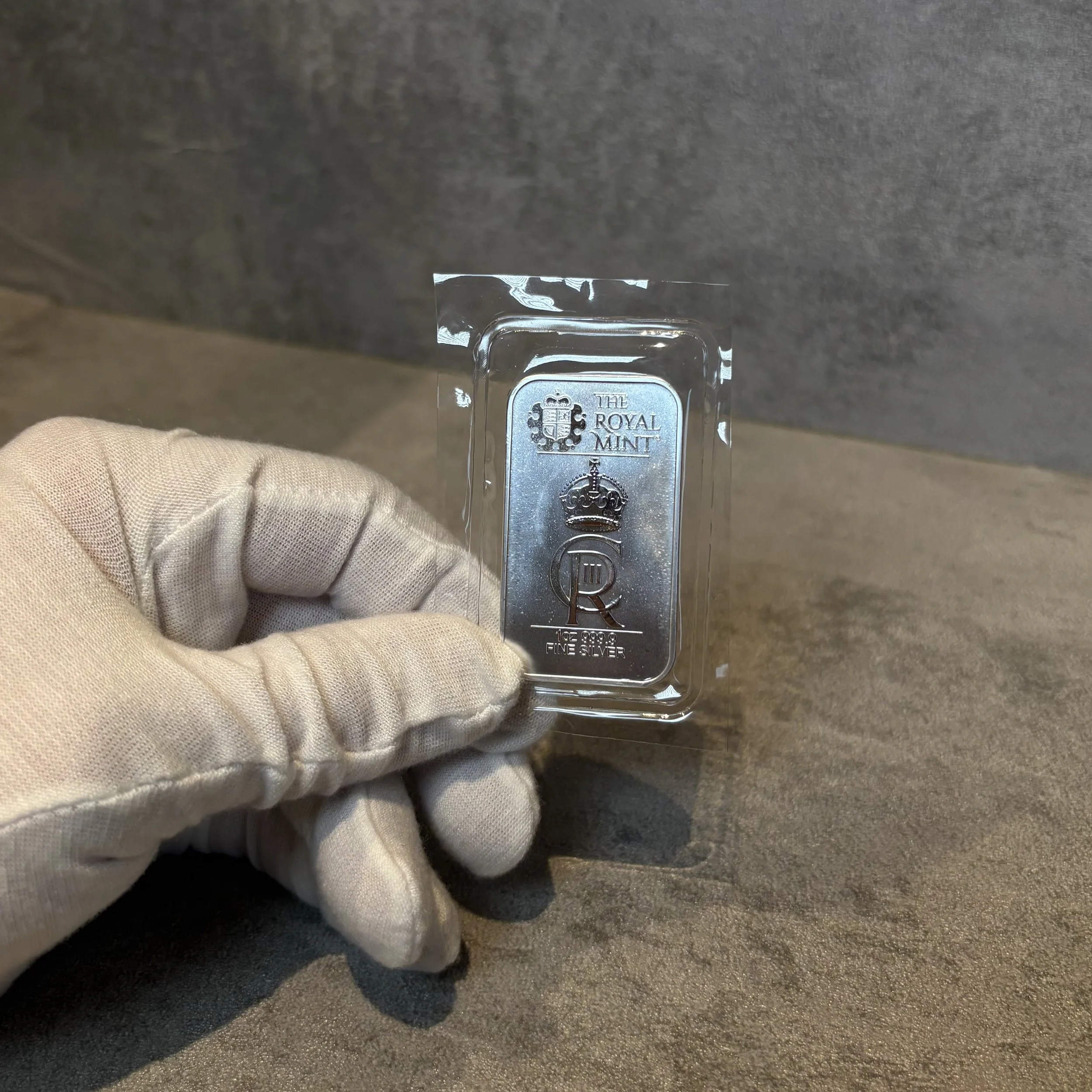 A gloved hand holding a plastic case with a silver bar engraved with the Royal Mint logo and the words 'The Royal Mint' and 'Fine Silver.'