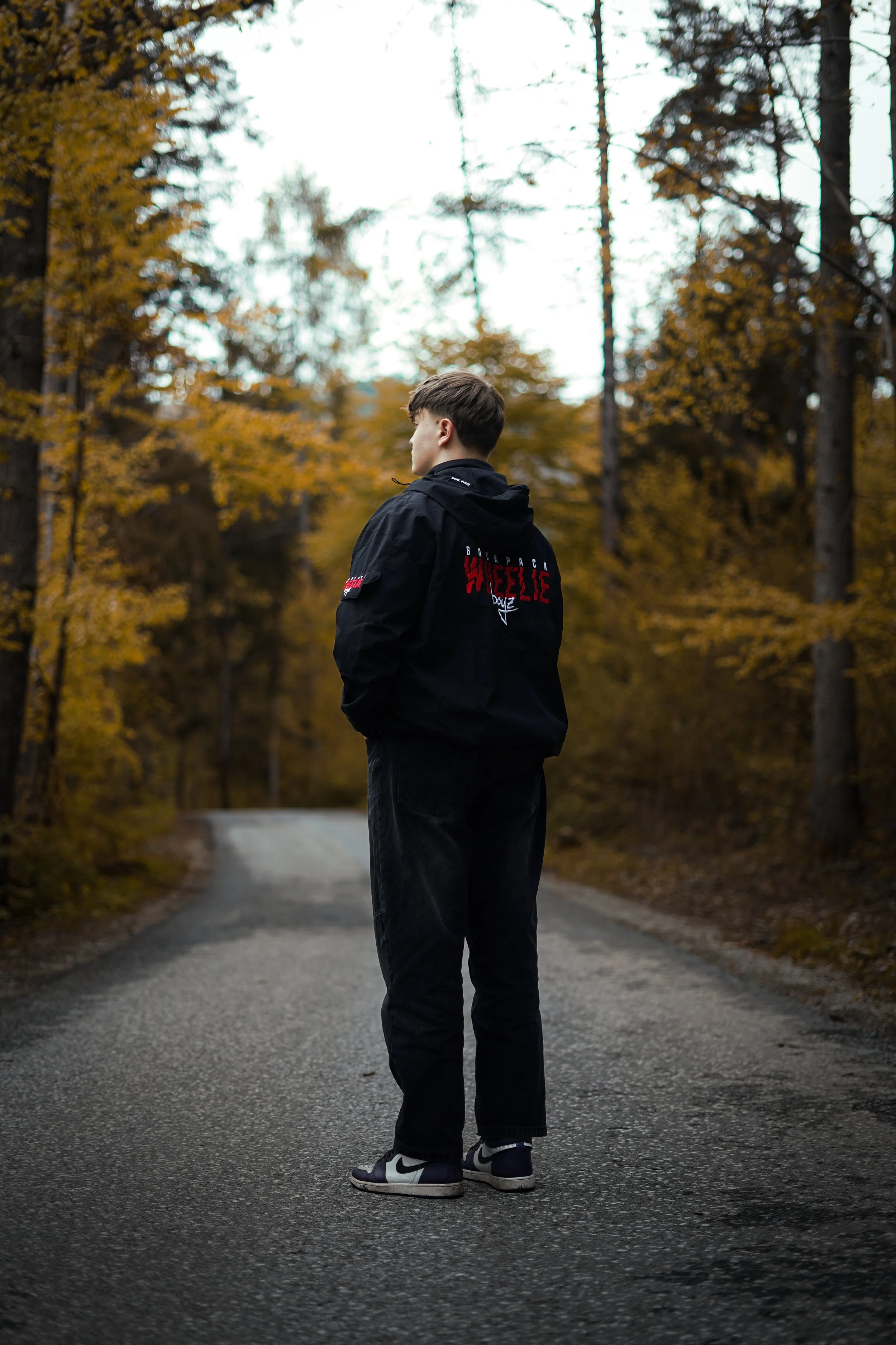 A young man in black clothing standing alone on a paved forest road with autumn trees around him, looking away from the camera.