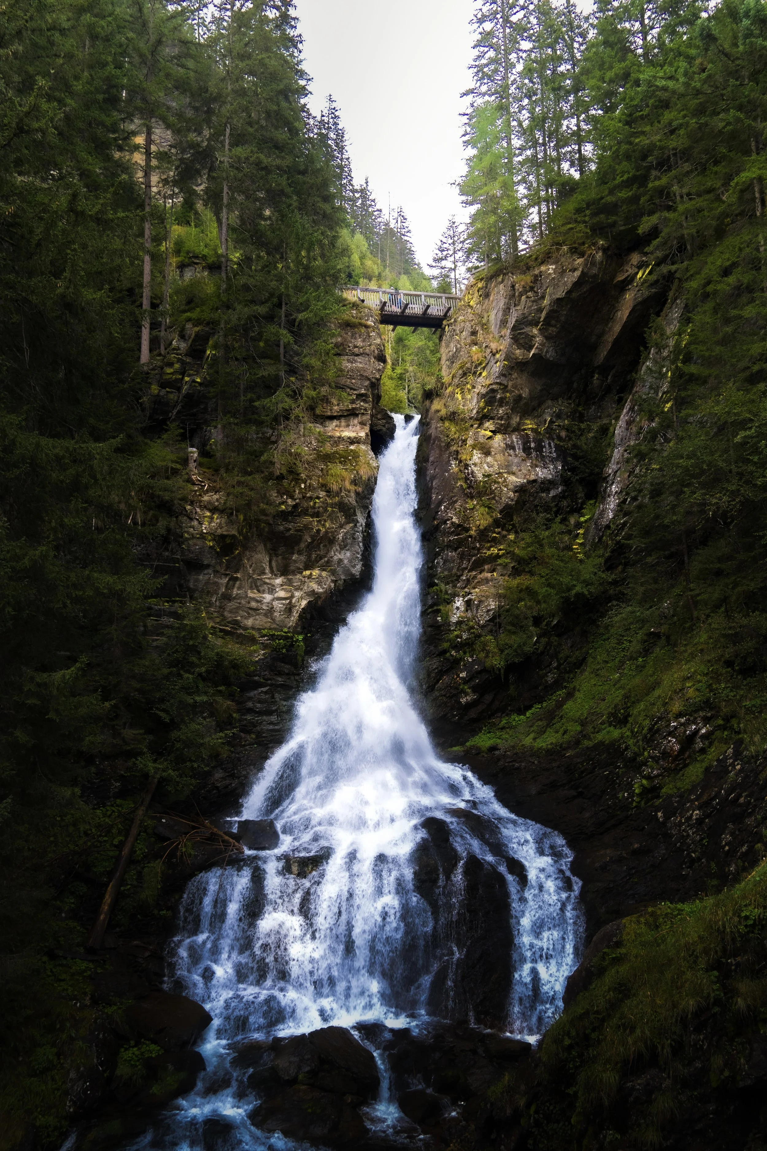 A waterfall cascading down a rocky cliff surrounded by lush green trees, with a wooden bridge at the top.