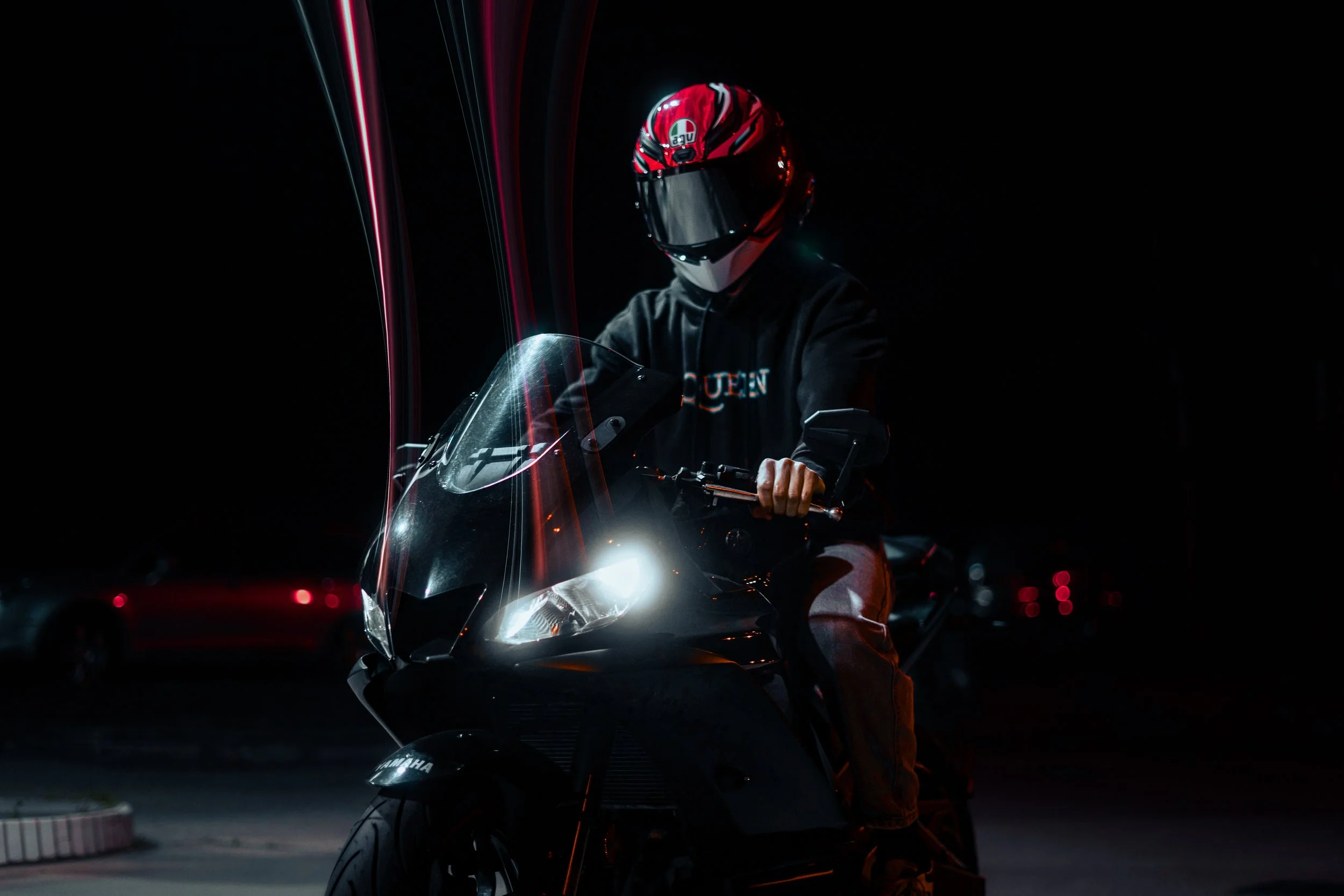 Person wearing a red and black motorcycle helmet and black hoodie, sitting on a black motorcycle with headlights on, in a dark setting with faintly visible background cars.