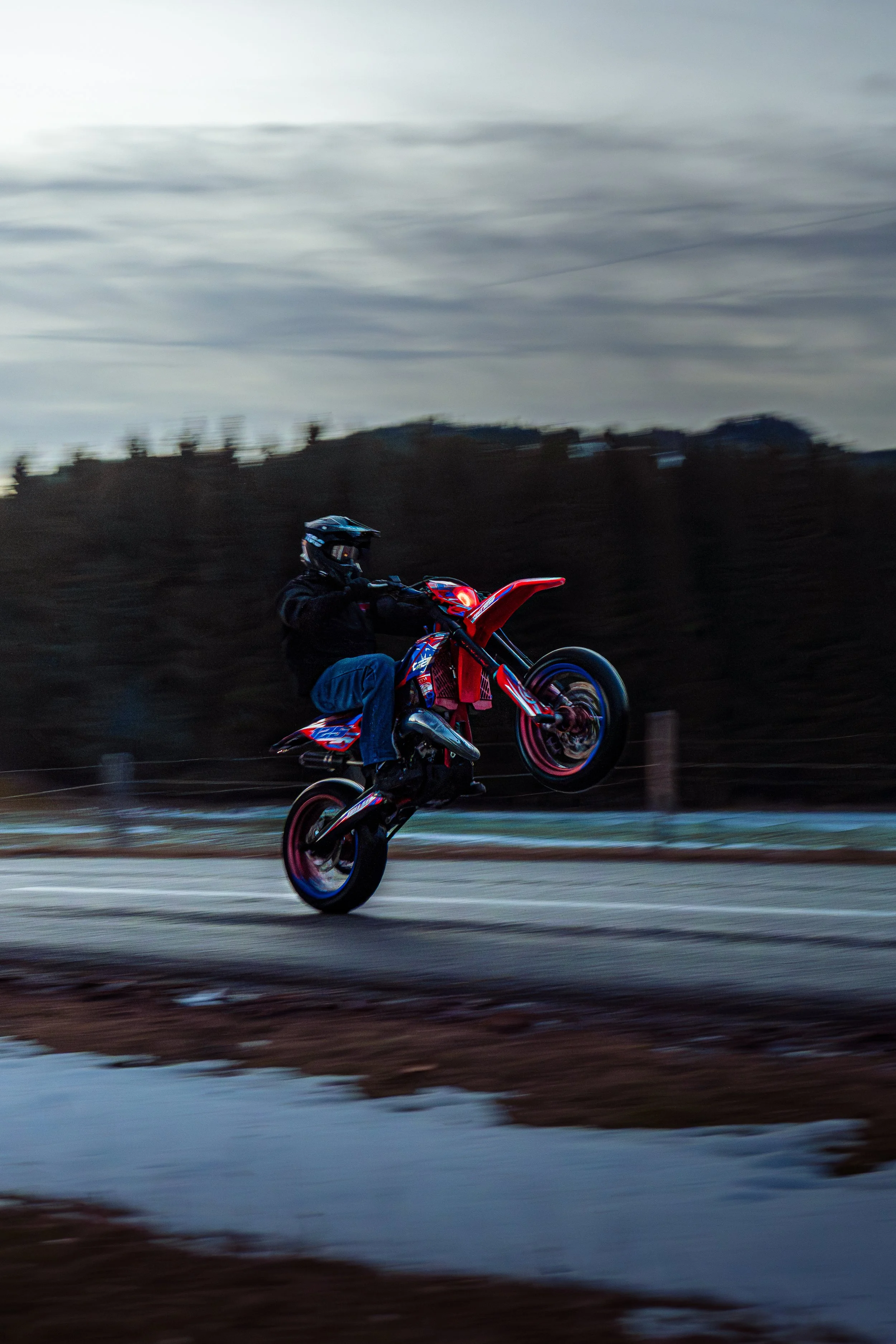 Person performing a wheelie stunt on a red dirt bike during dusk or dawn, with a blurred background of trees and cloudy sky.