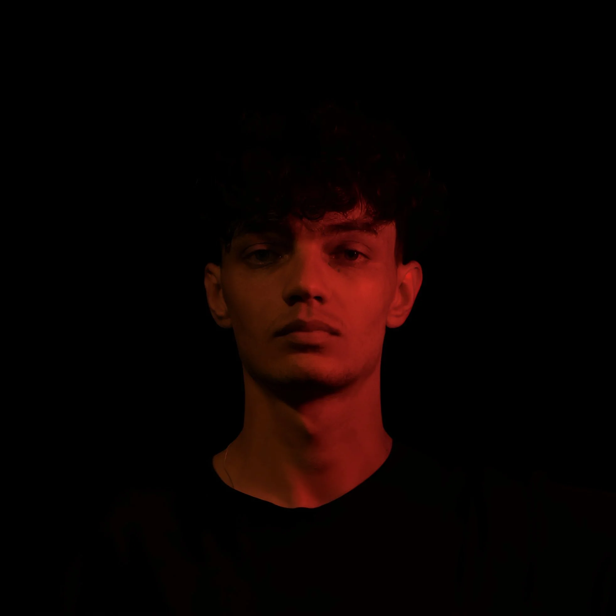 Close-up portrait of a young man with curly hair, illuminated with red lighting against a black background.