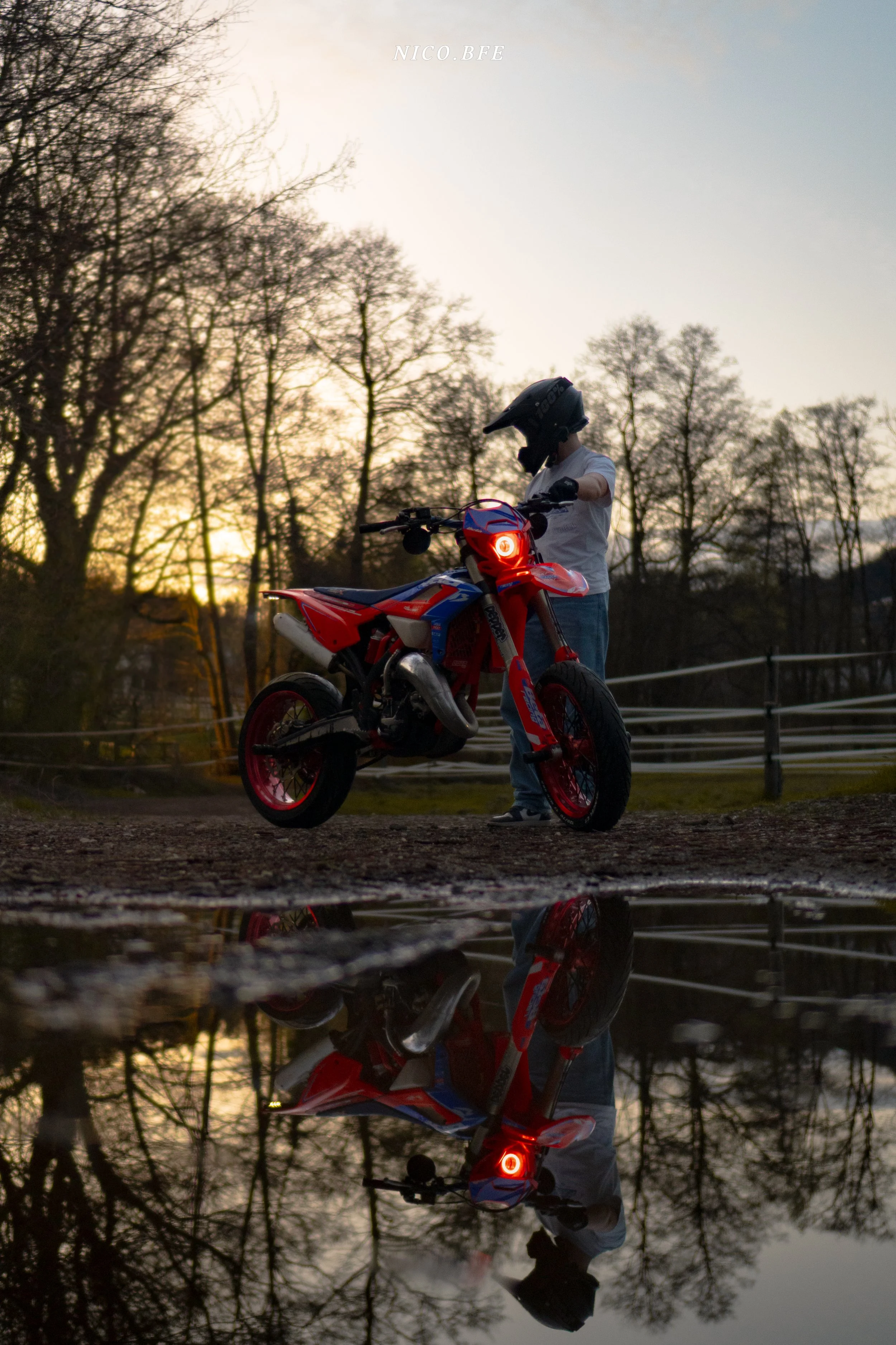 Person wearing a helmet standing next to a red and blue dirt bike near a puddle at sunset, with trees in the background.