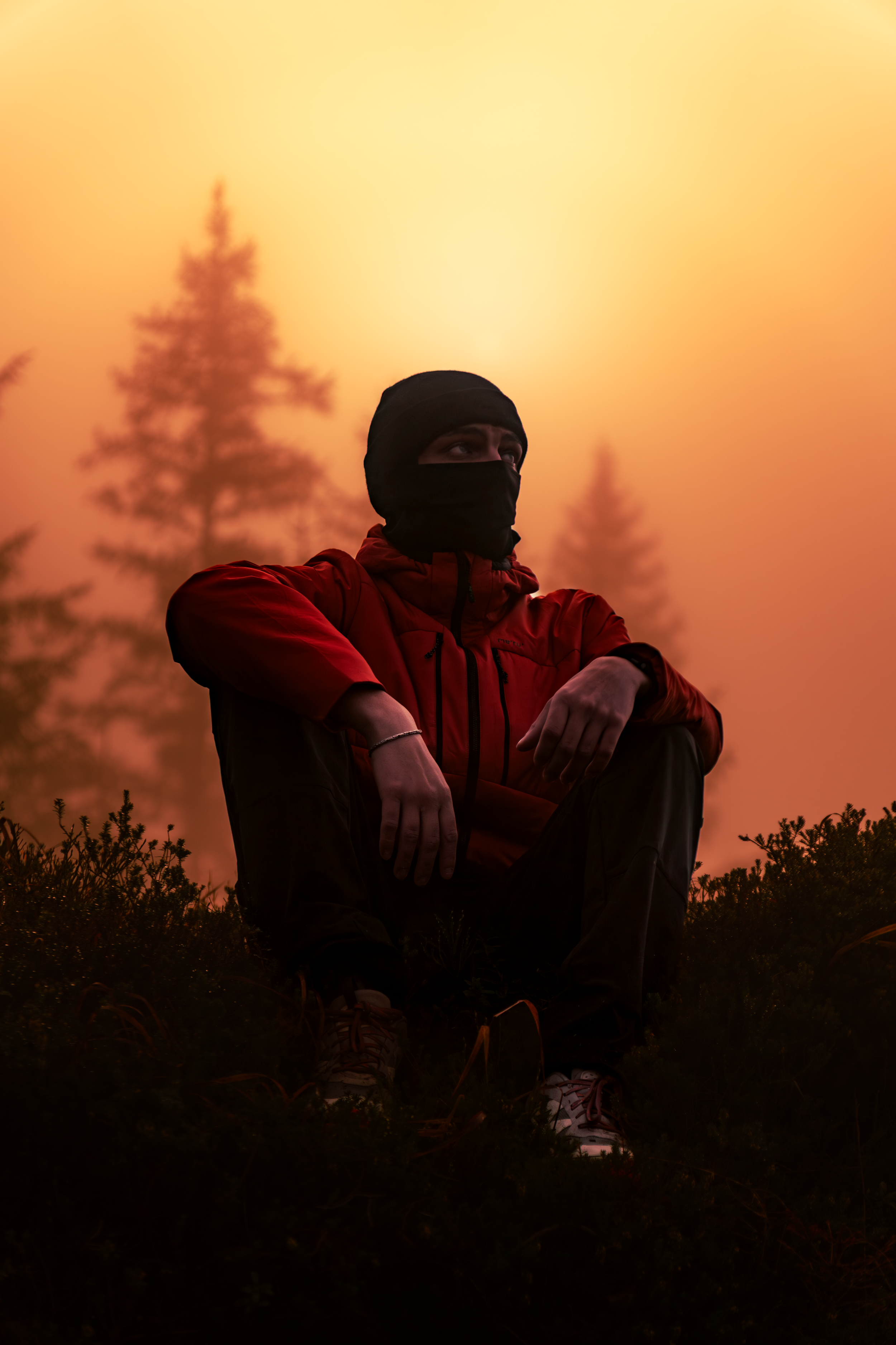 Person sitting outdoors during sunset, wearing a red jacket and black face covering, with trees in the background.