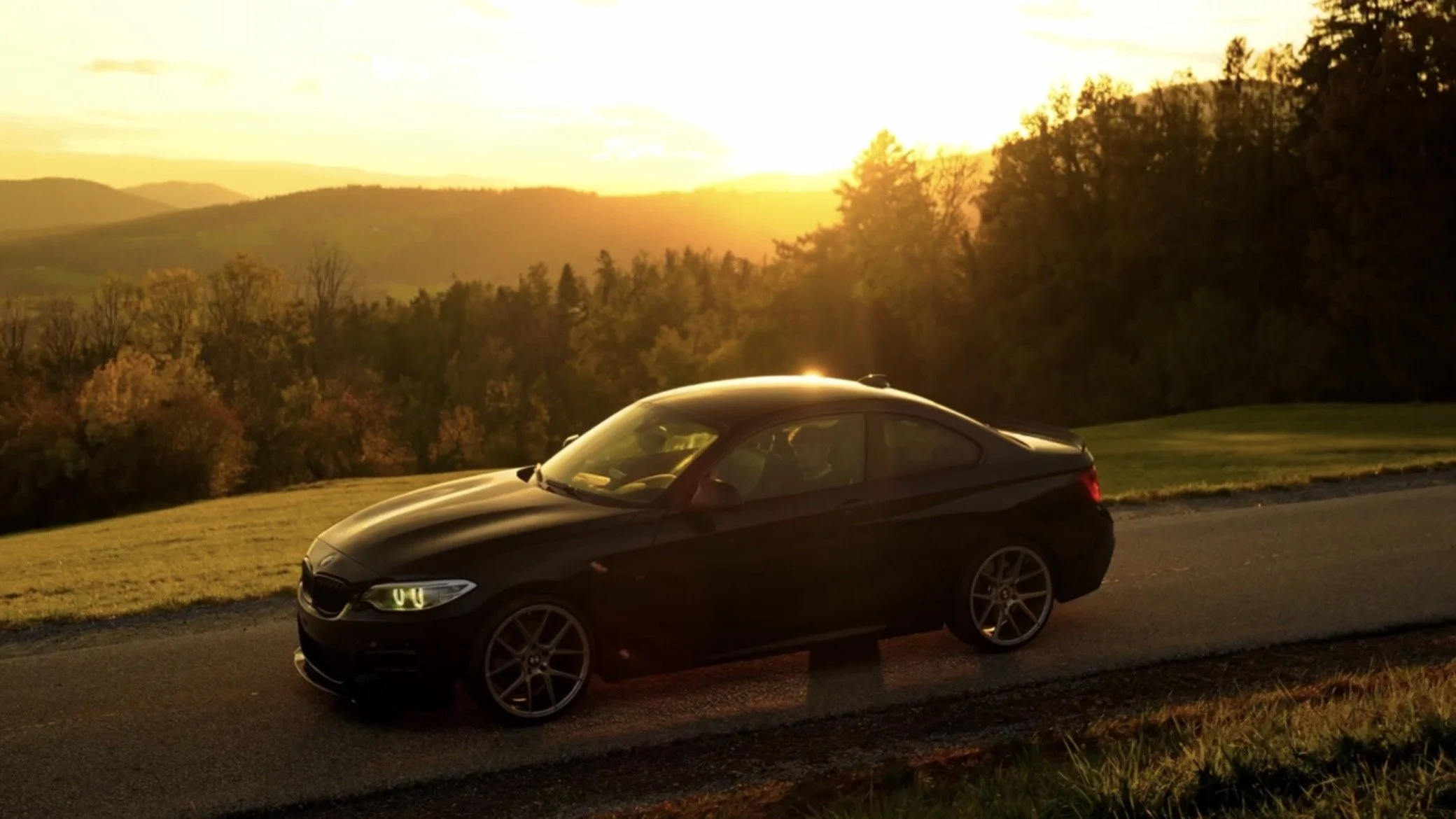Black coupe car parked on a roadside with a scenic sunset in the background, hills, and trees.