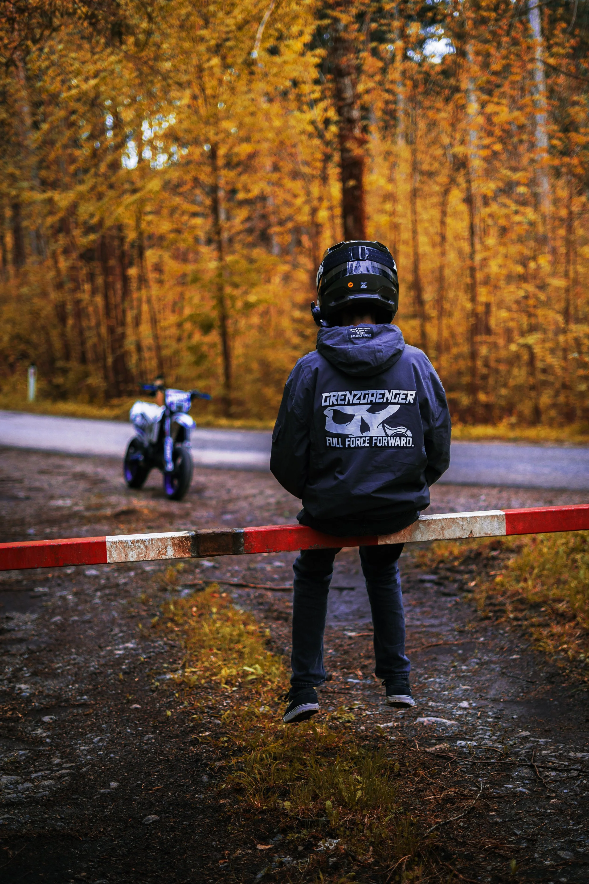 A person wearing a black helmet and a dark jacket with a skull logo, sitting on a red and white barrier in a forest with autumn-colored trees. A motorcycle is in the background on the dirt road.