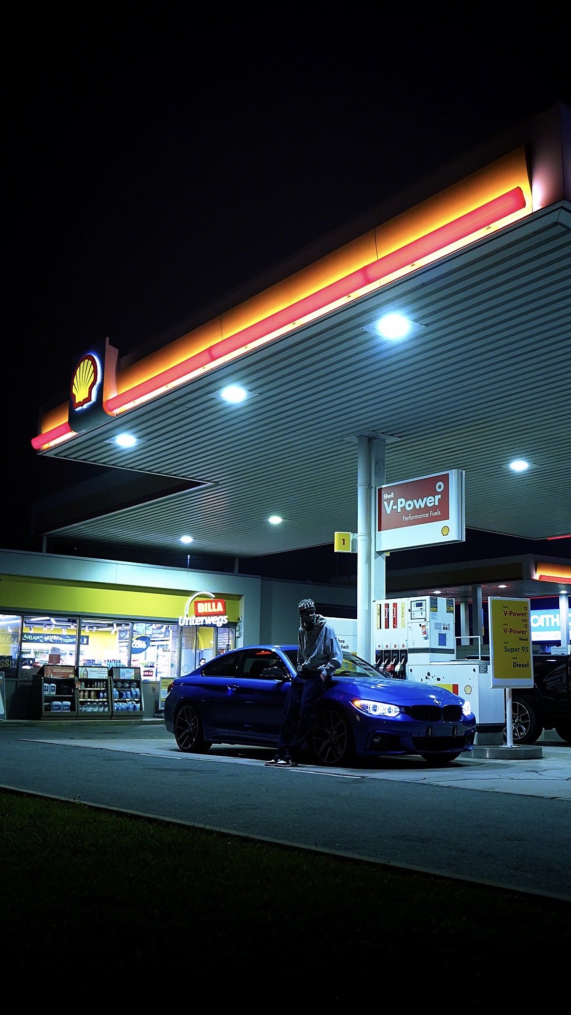 A man in a hoodie and cap standing next to a blue car at a Shell gas station at night, with illuminated signs and a convenience store in the background.