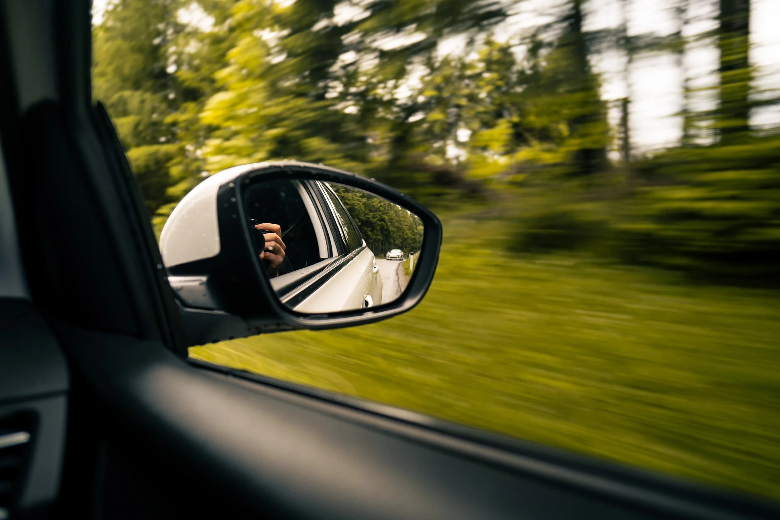 View from inside a car looking into the side mirror, showing a road behind with trees on both sides and a white car approaching in the distance.