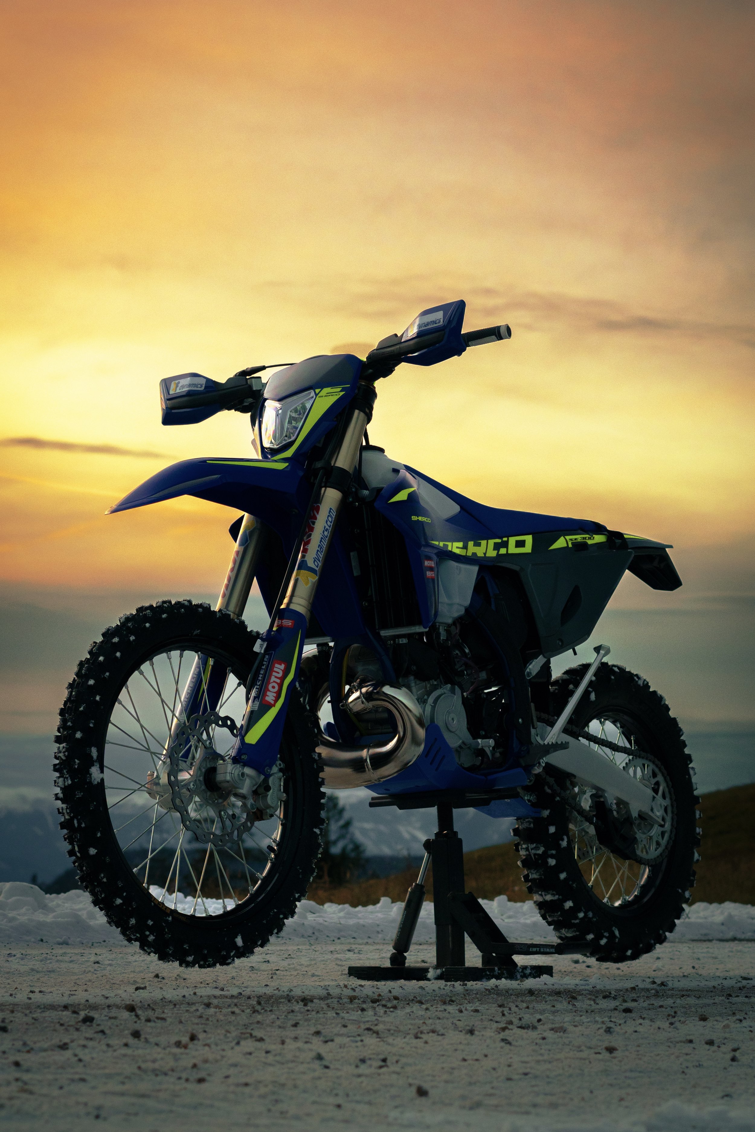 A dirt bike parked on a snowy ground during sunset with a mountain landscape in the background.