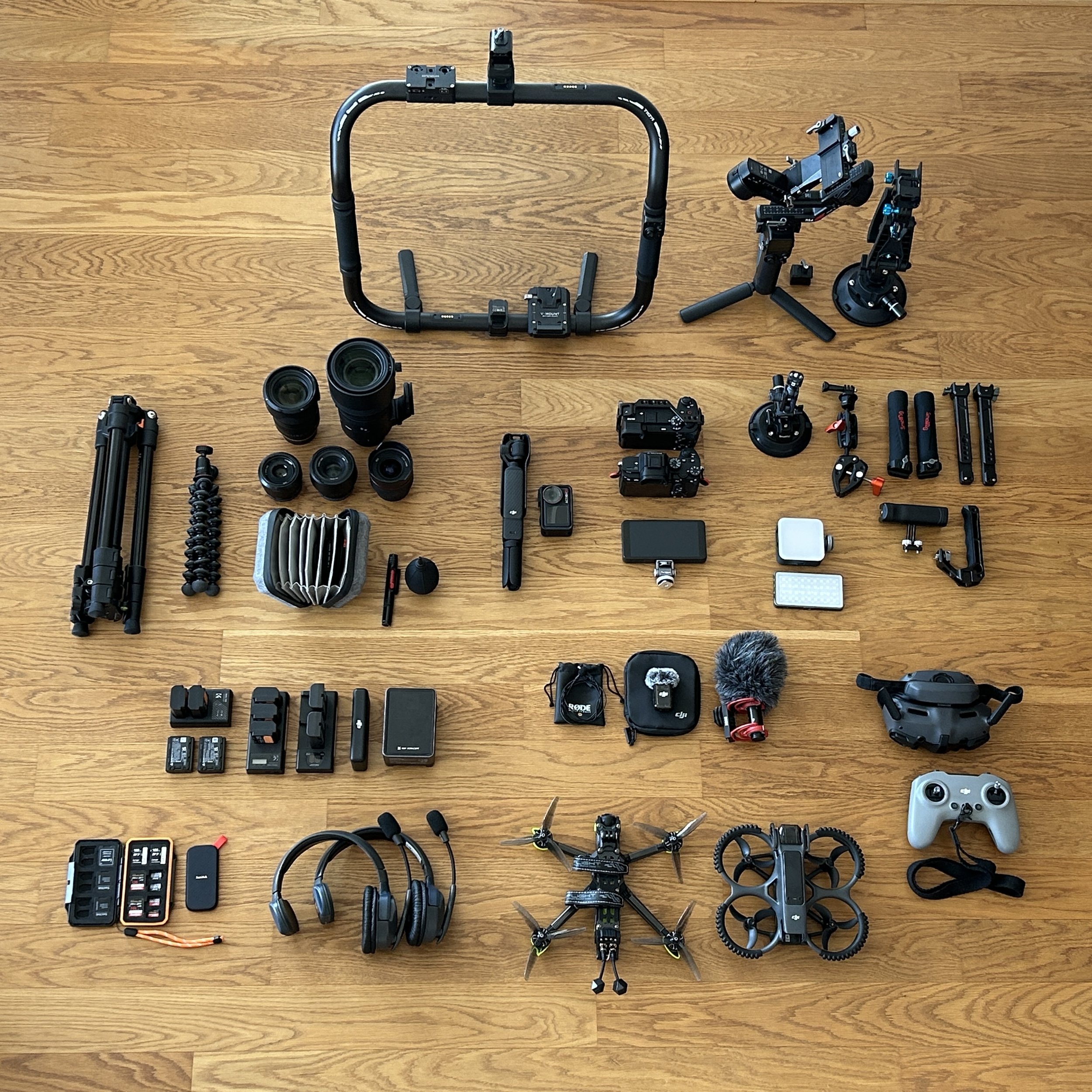 A variety of professional camera equipment, including cameras, lenses, tripods, stabilizers, drones, microphones, controllers, and accessories, all arranged on a wooden floor.