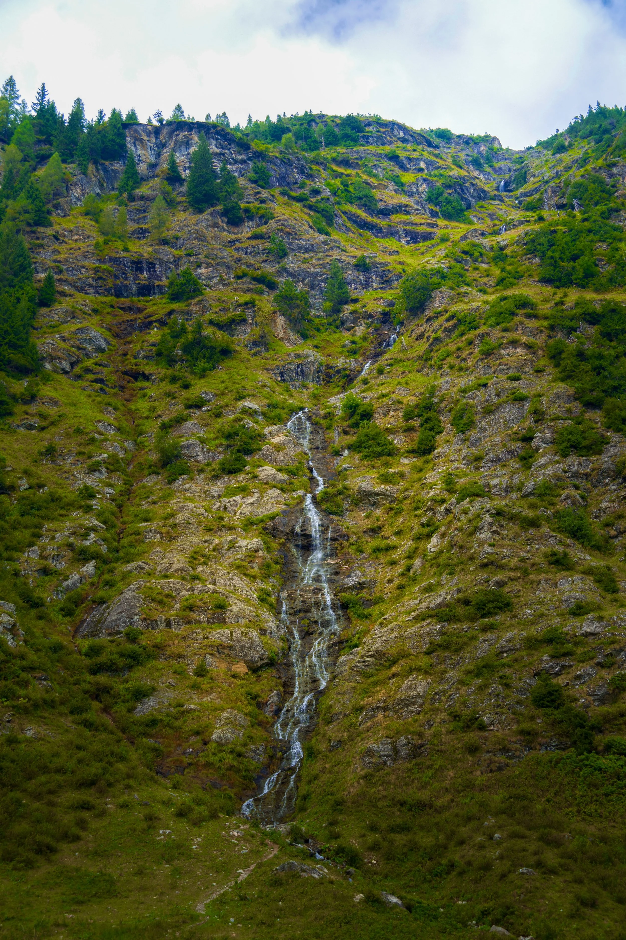 A tall mountain with a small waterfall flowing down a rocky and grassy slope, surrounded by green vegetation and trees at the top.