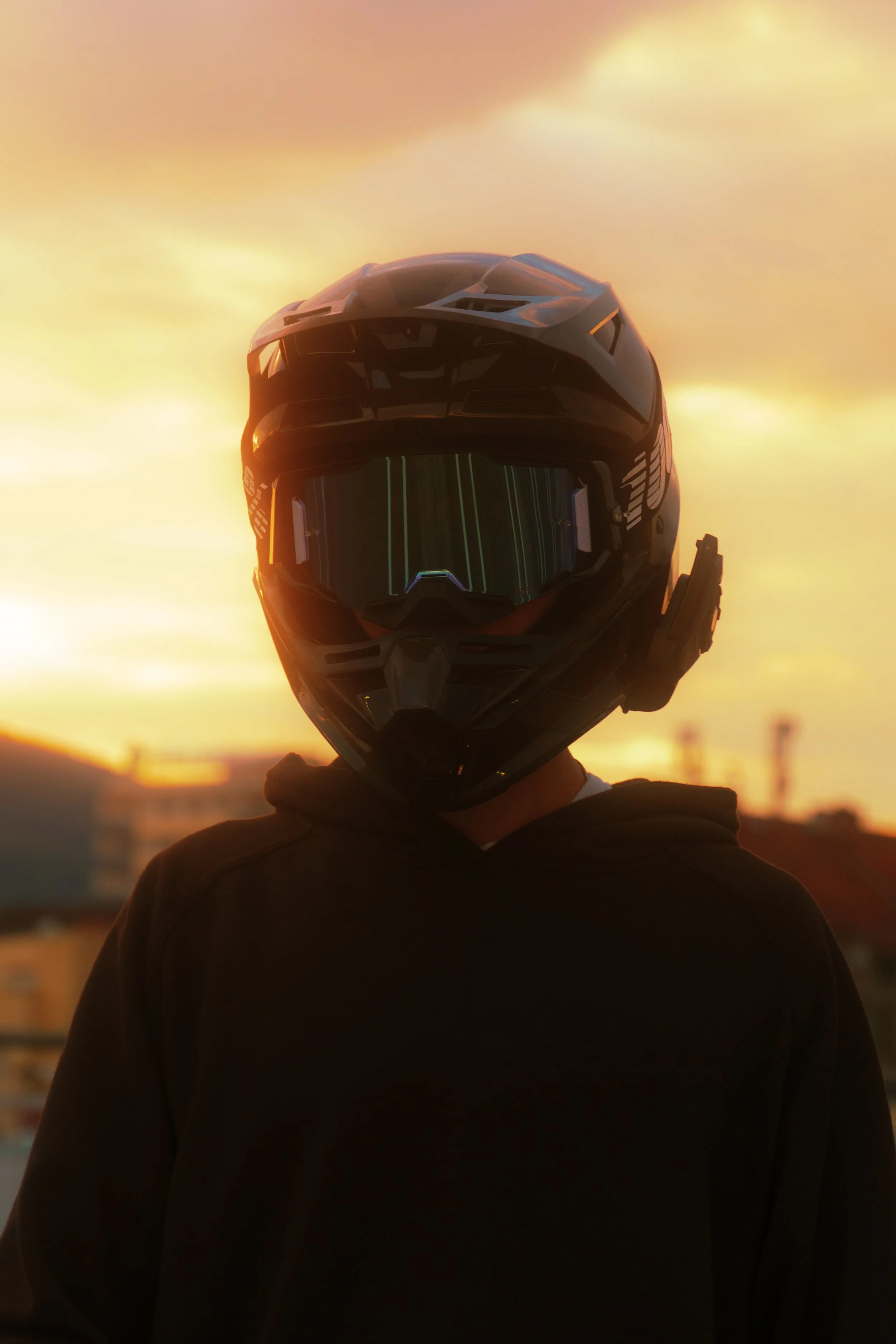 Person wearing a black motorcycle helmet and hoodie, standing outdoors during sunset with colorful sky in the background.