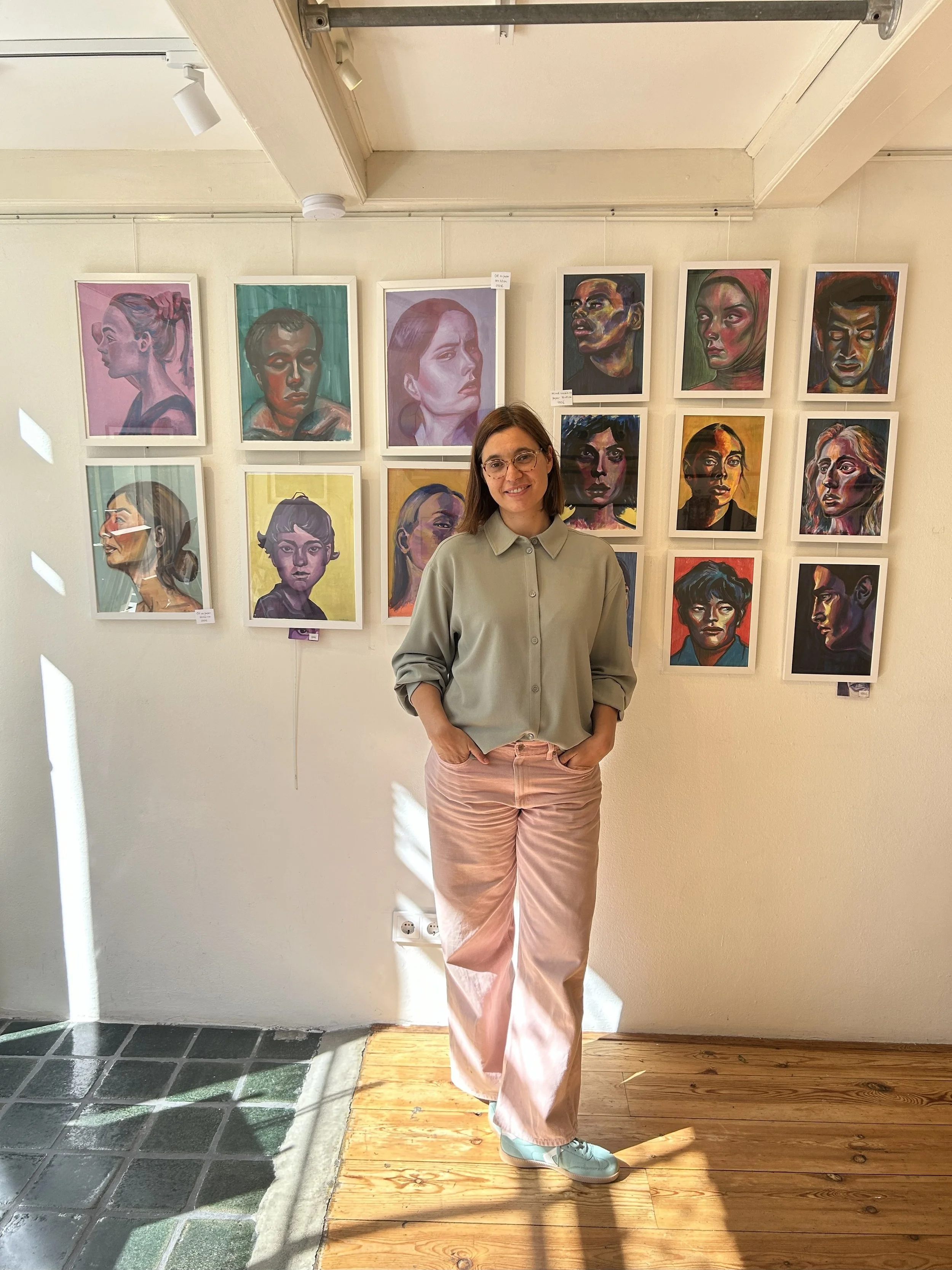A woman with glasses, brown hair, wearing a green button-up shirt, pink pants, and blue shoes, standing in front of a wall with colorful portraits at an art gallery.