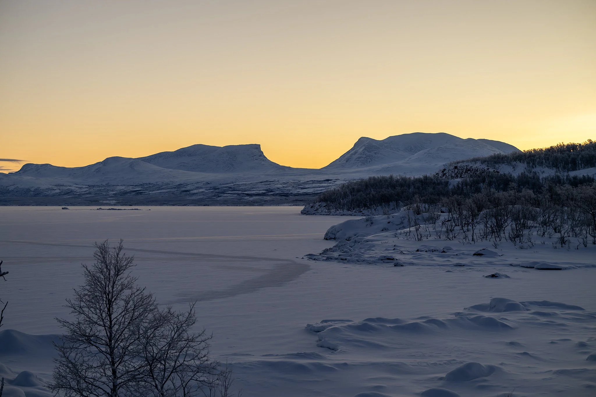 Snow-covered landscape with leafless trees in foreground, frozen lake or river in the middle, and mountains in the background during sunset or sunrise.