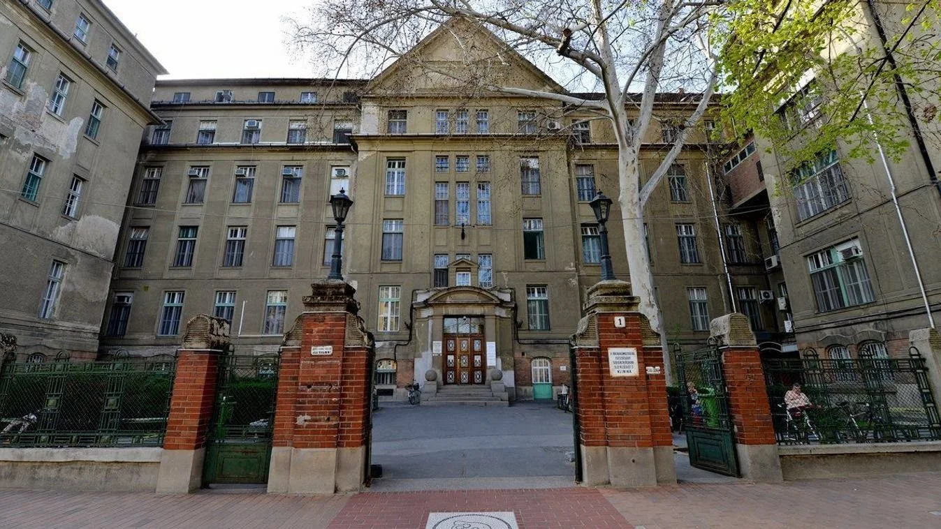 Old multi-story building with a gated entrance, brick pillars, and benches outside. Trees and street lamps are also visible.