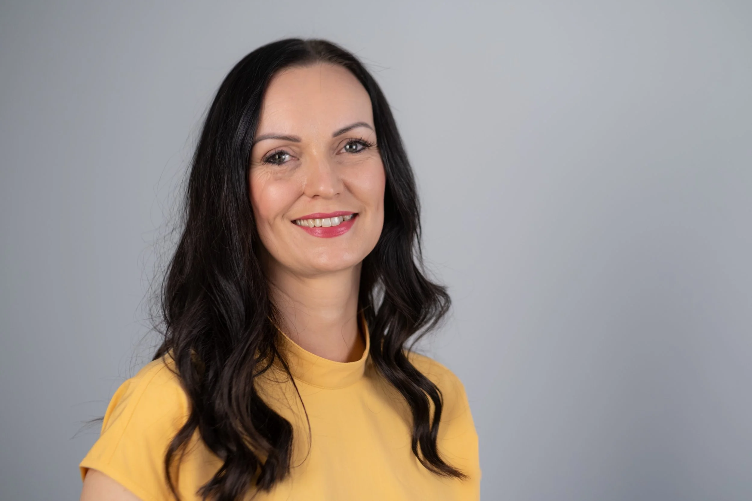 A woman with long wavy dark hair wearing a yellow top, smiling, against a plain light gray background.