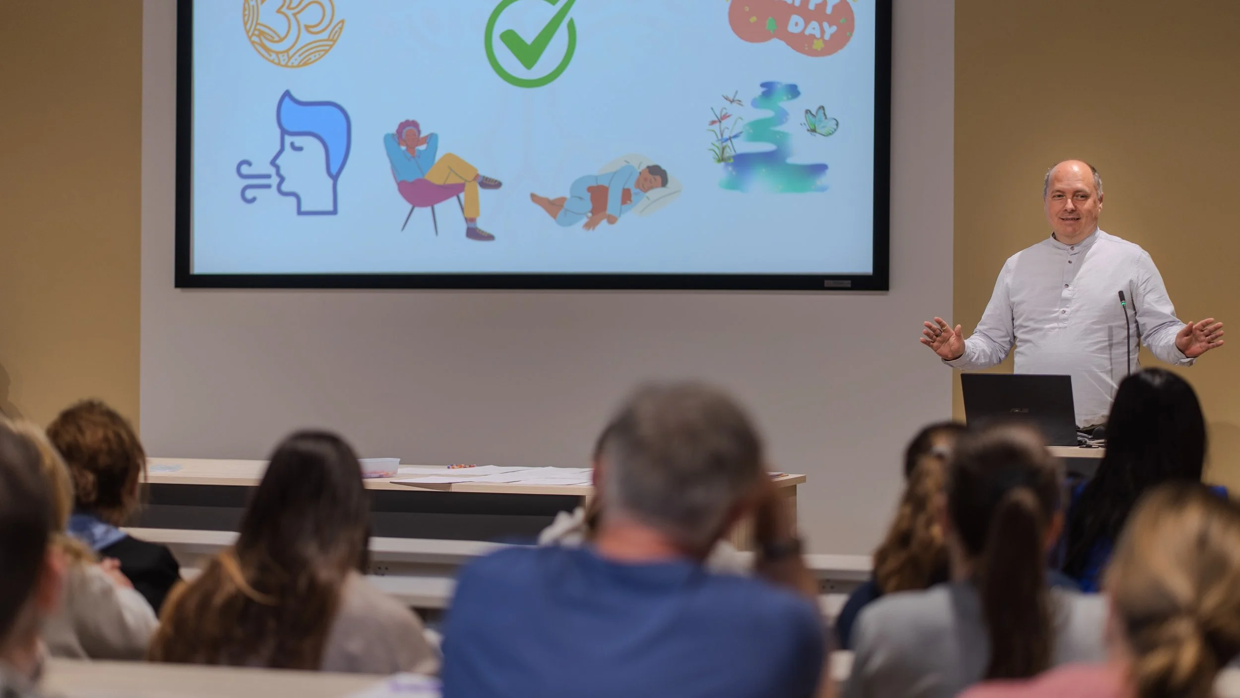 A man in a white shirt gives a presentation to an audience in a classroom or conference room. There is a large screen behind him displaying various colorful graphics and icons, including a check mark, a person sleeping, and other illustrations.