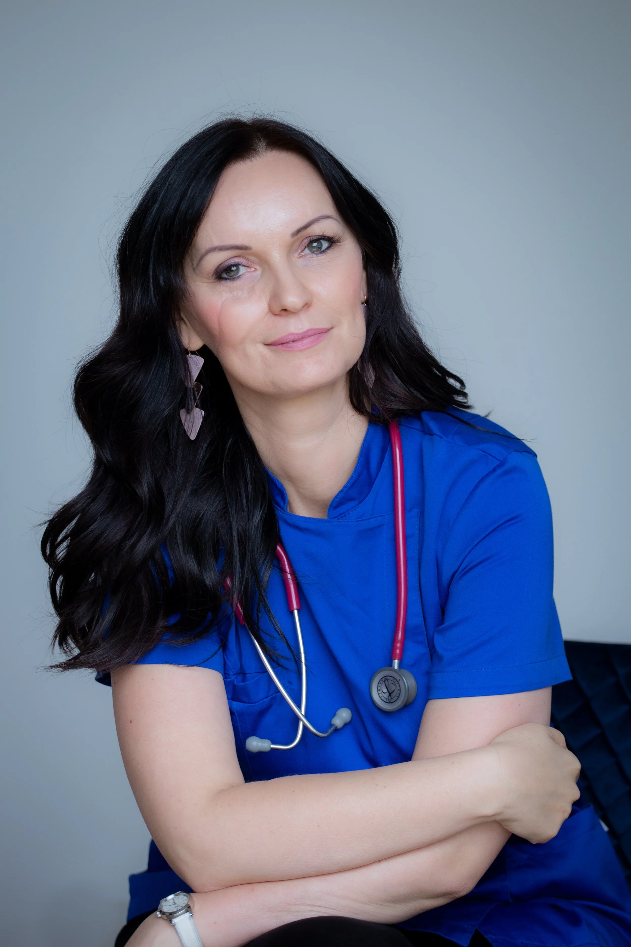 A woman with long dark hair wearing blue medical scrubs and a stethoscope around her neck.