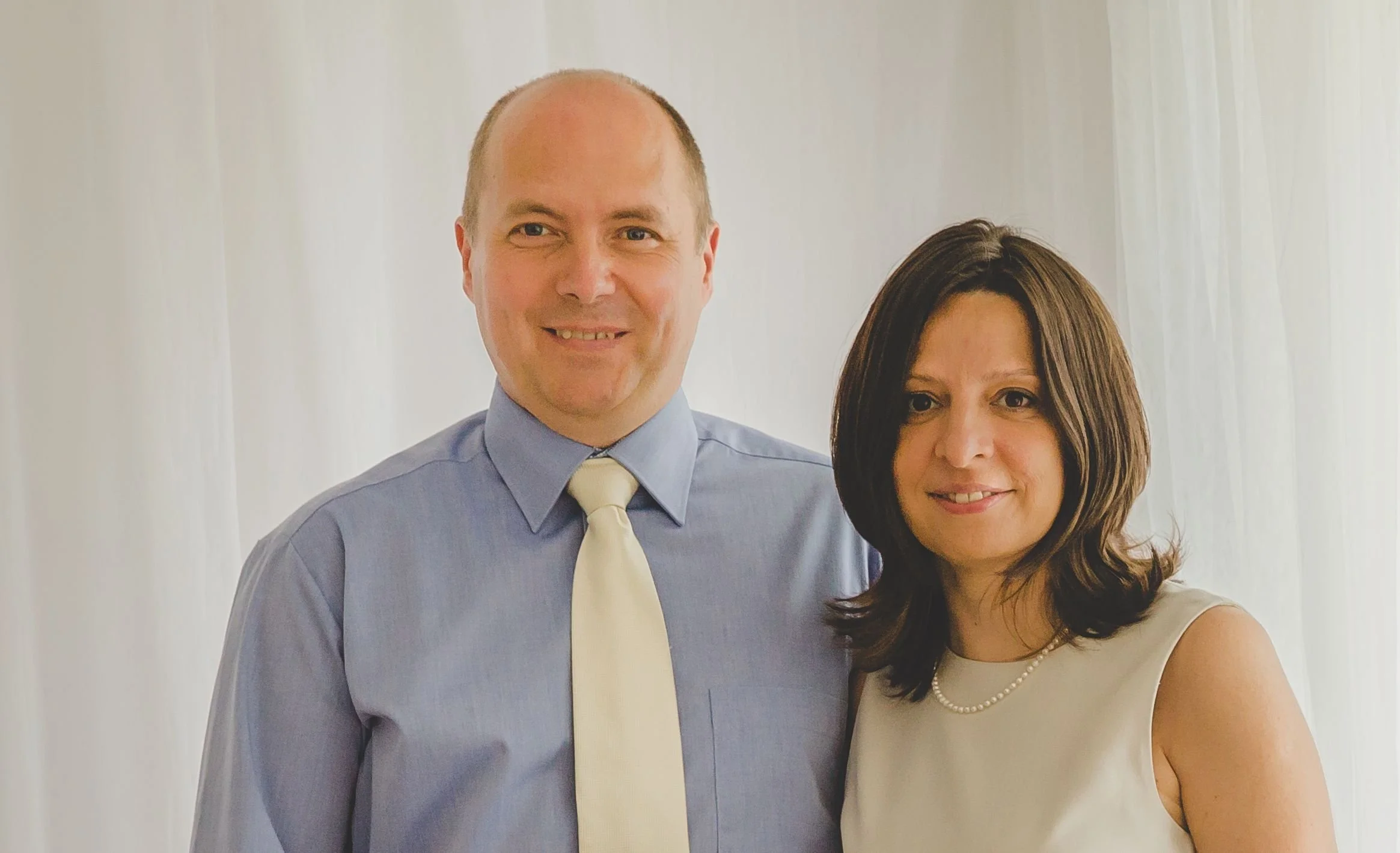 A man and woman standing together, smiling, in front of white curtains.
