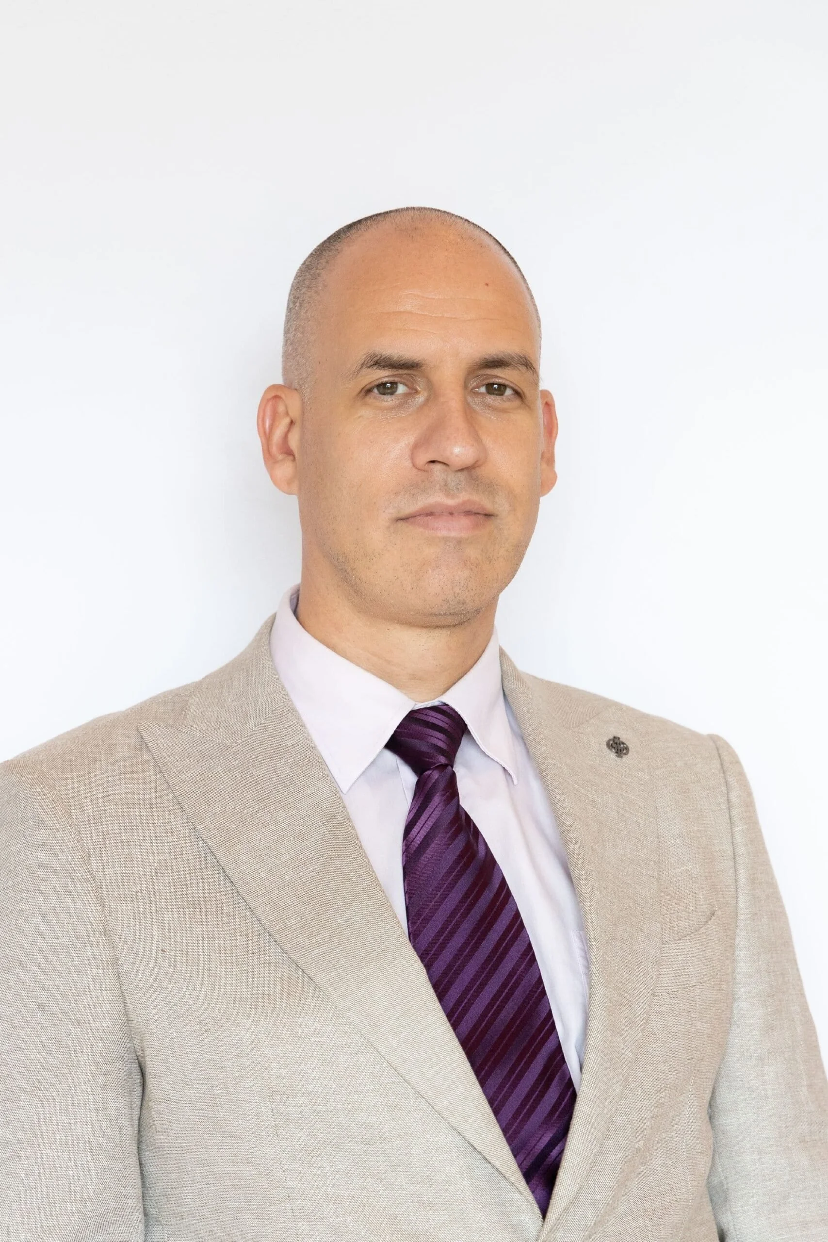 A man in a beige suit with a purple striped tie poses against a plain white background.