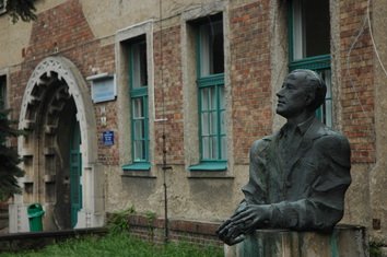 Statue of a man in front of a brick building with green window frames and an arched doorway.
