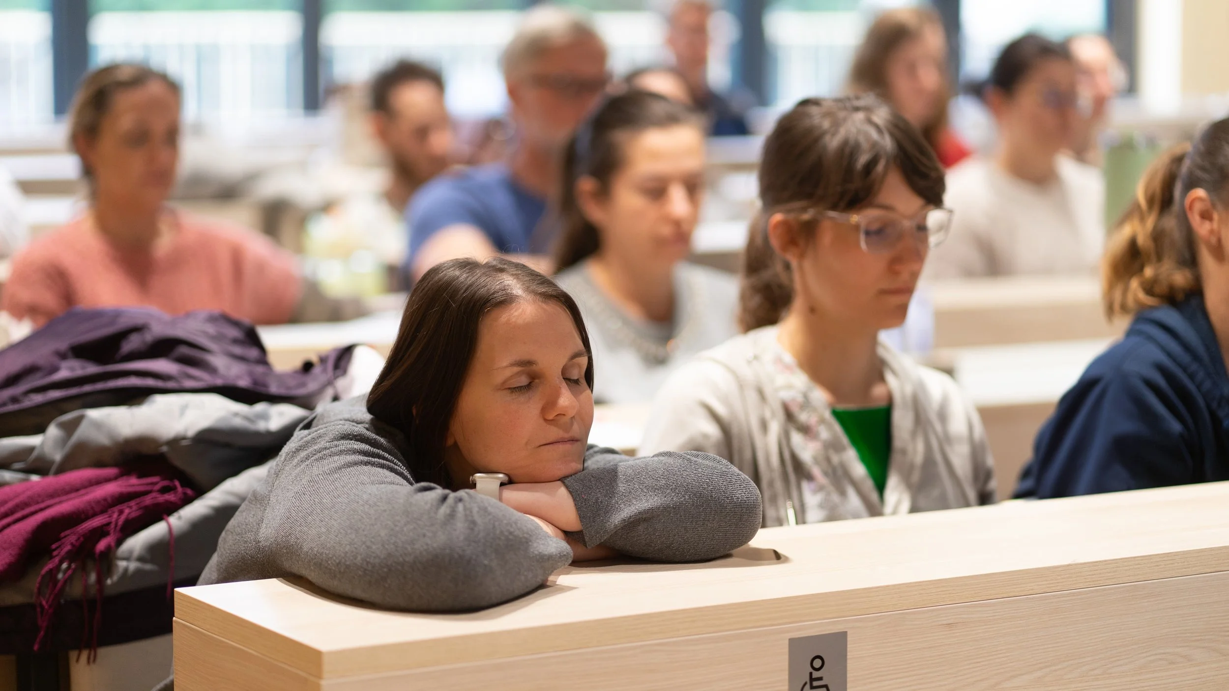 A woman resting her head on her arms with eyes closed in a classroom with other students sitting at desks, some with eyes closed, possibly sleeping or meditating.