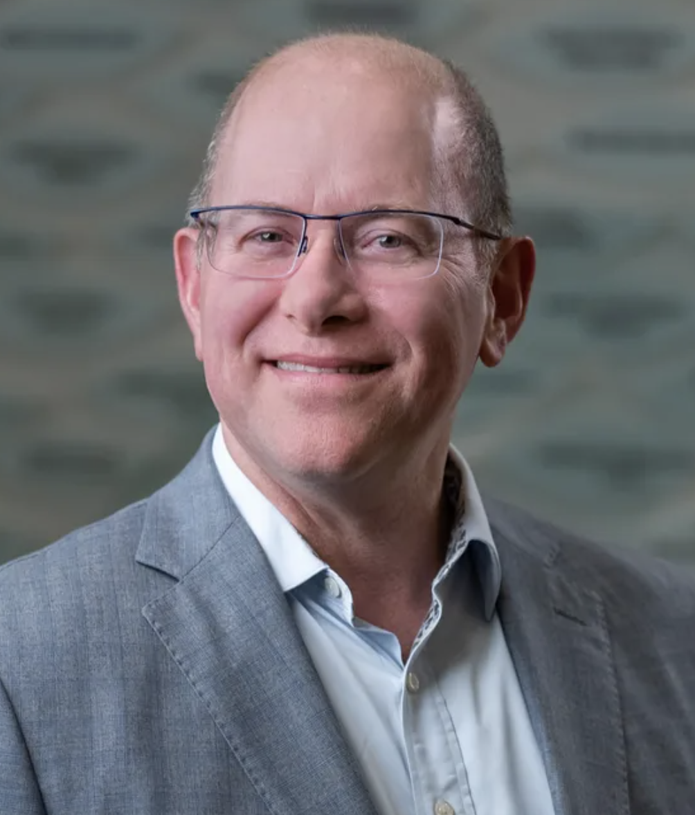 Professional portrait of a middle-aged man with glasses, wearing a gray suit and a light-colored shirt, smiling against a blurred gray background.