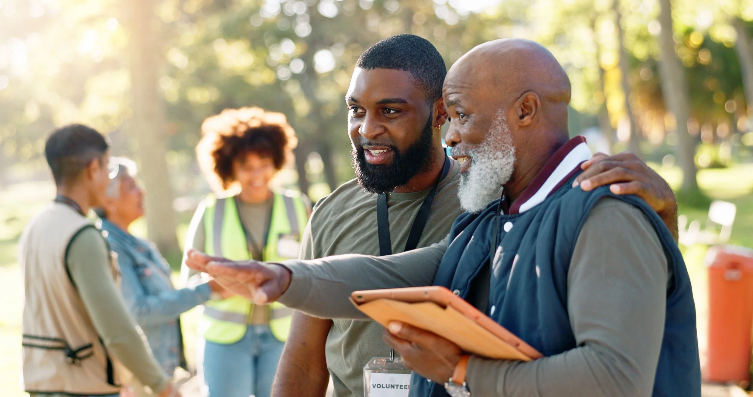 Two men talking and smiling at an outdoor event, with others in the background and trees around.
