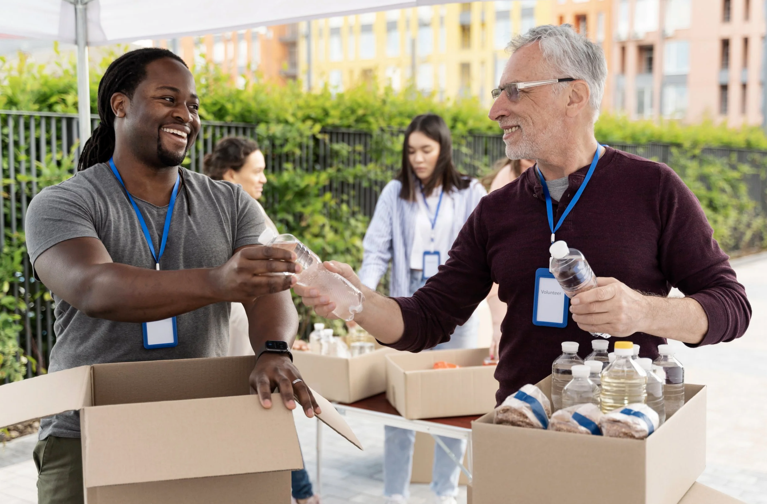 Two men smiling and exchanging a bottle of water at an outdoor volunteer event with boxes of food and bottles on a table, with others in the background and colorful buildings behind.