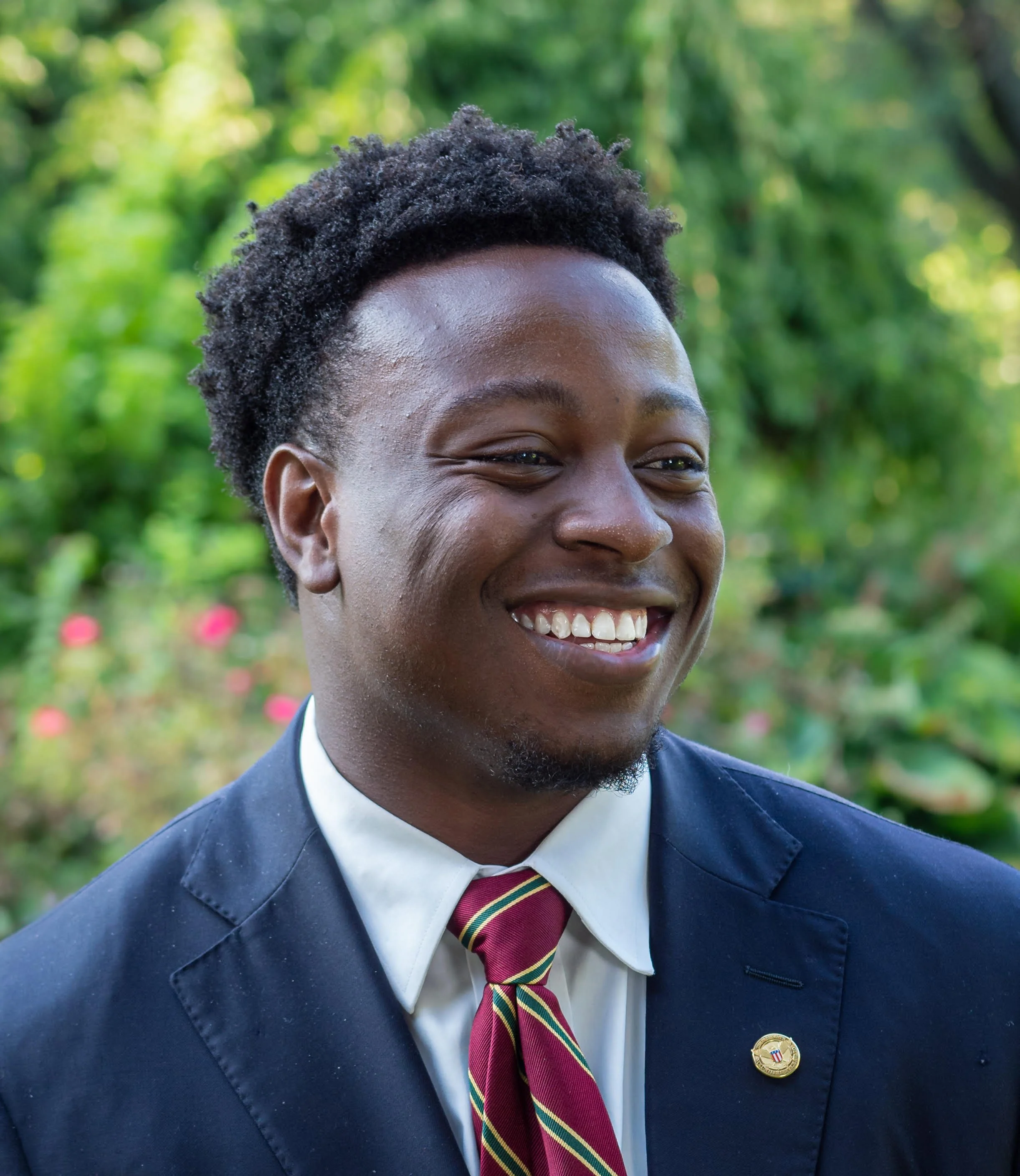 Close-up of a smiling young man with dark curly hair, wearing a suit, white shirt, and striped tie, with a pin on his lapel, outdoors with blurred green foliage in the background.