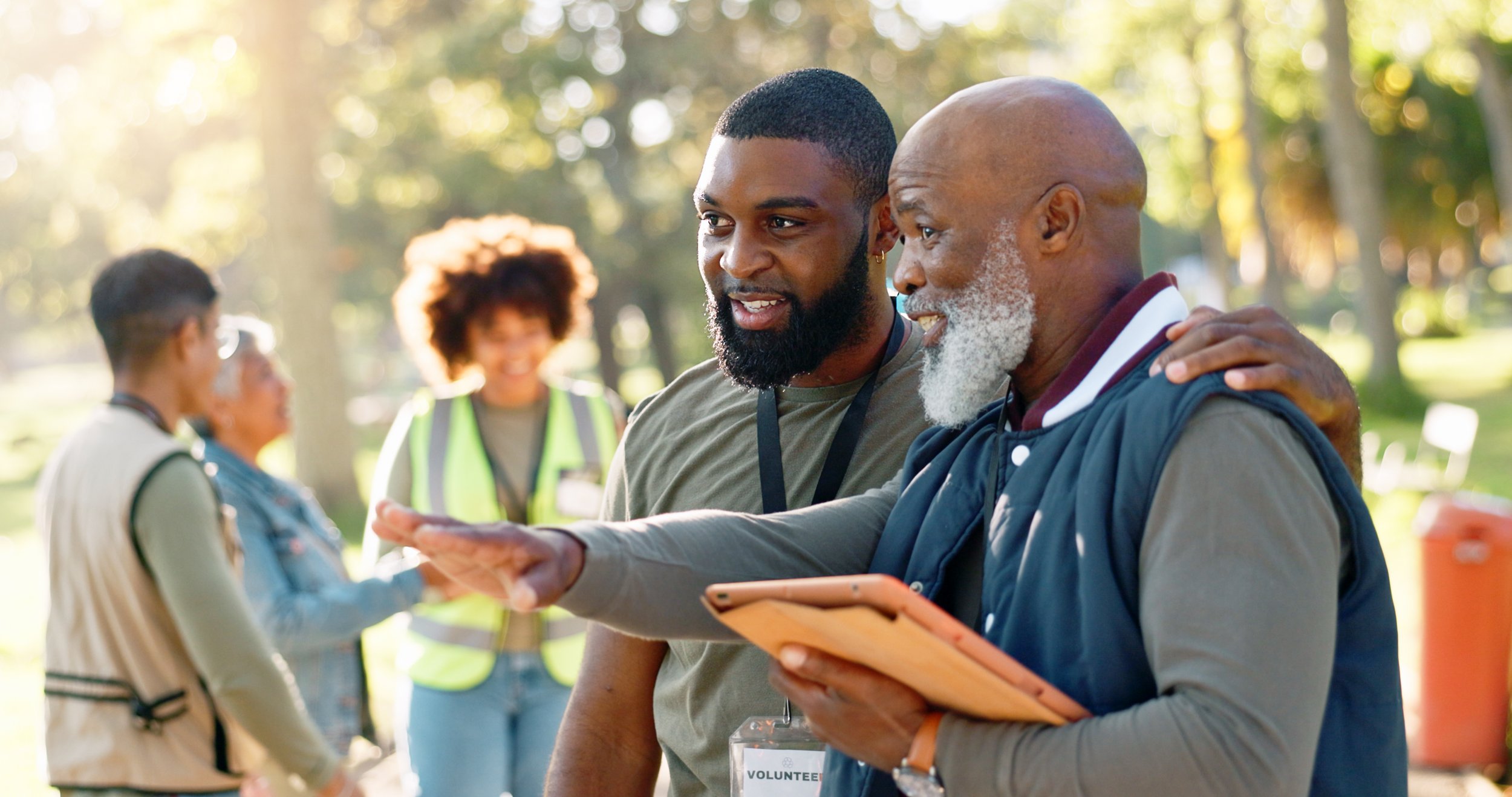 Two Black men, one with a beard, shaking hands at an outdoor volunteer event in a park with trees and sunlight, with two women in the background, one wearing a yellow vest and sunglasses, and the other with curly hair, all smiling and engaging in conversation.