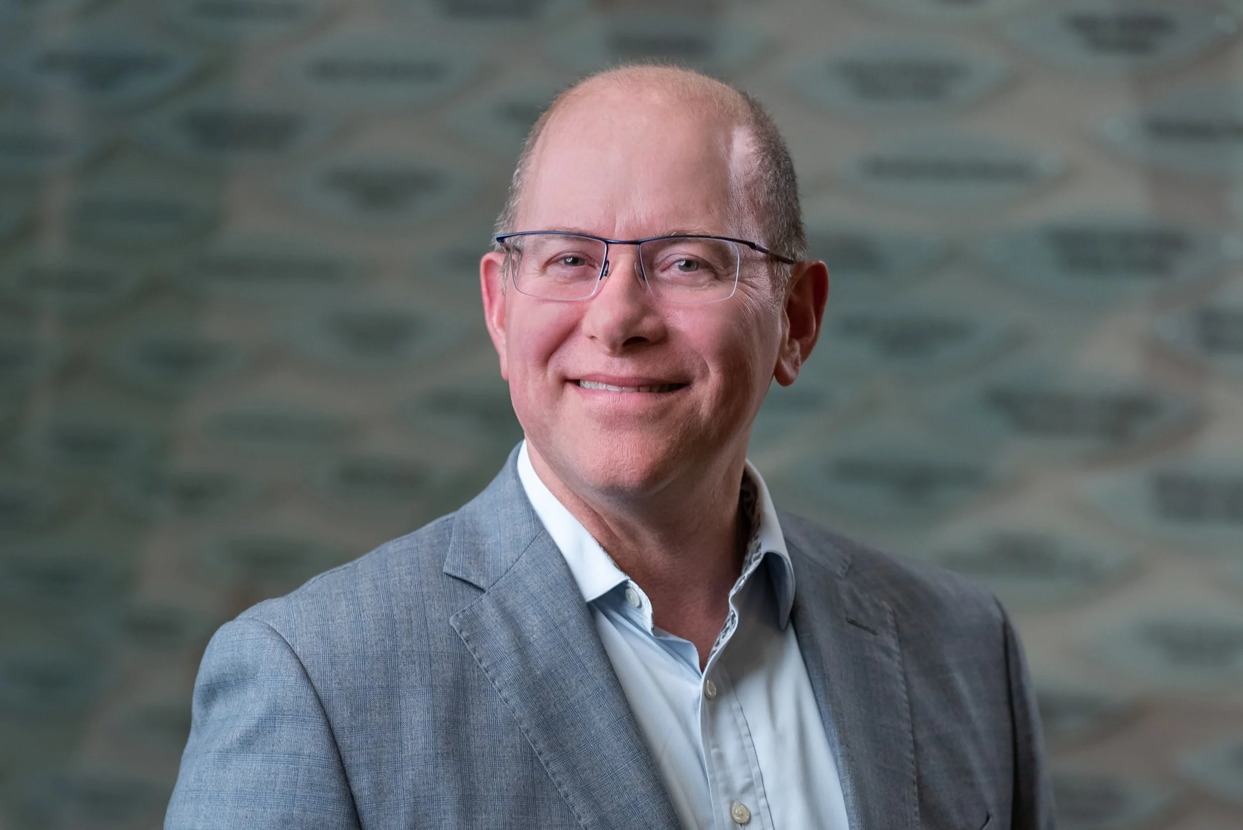 Close-up portrait of a middle-aged man with glasses, wearing a grey suit jacket and white shirt, smiling, standing in front of an abstract background.