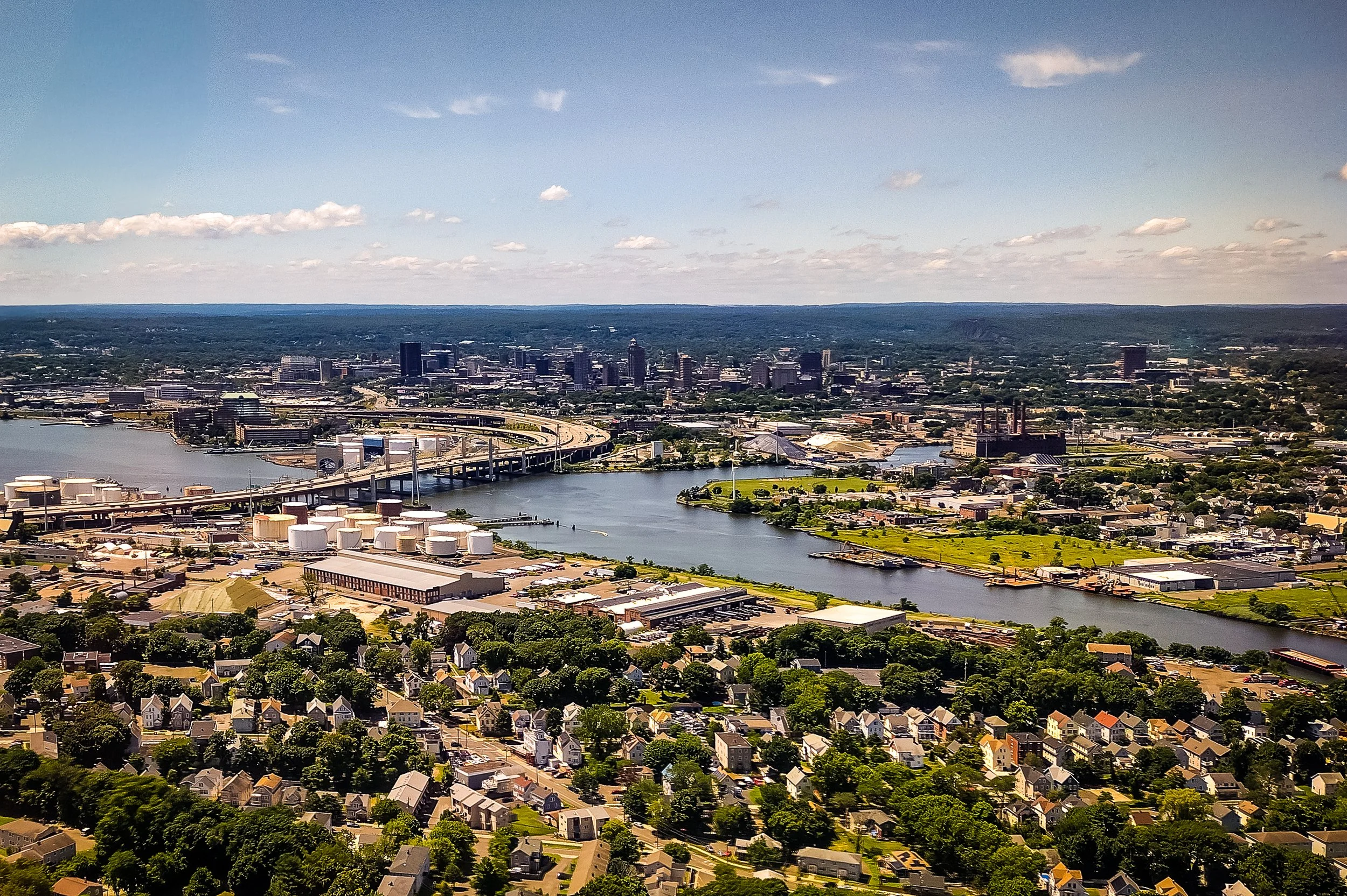 Aerial view of a city skyline with tall buildings, a river, bridges, and residential neighborhoods under a partly cloudy sky.