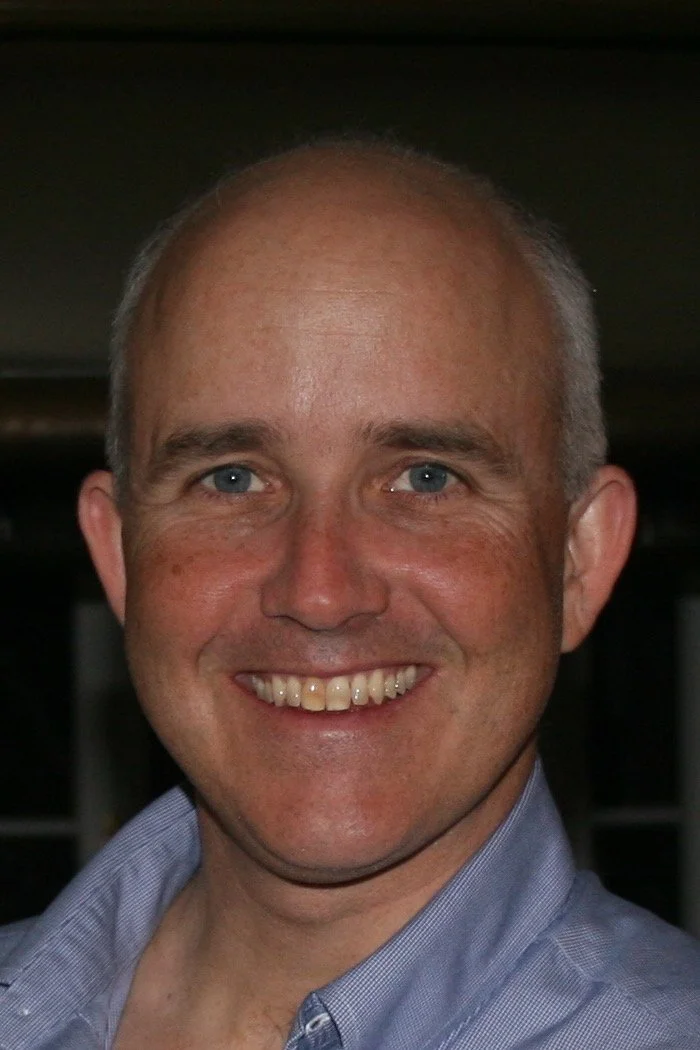 Close-up of a smiling man with light skin and blue eyes, wearing a blue collared shirt.