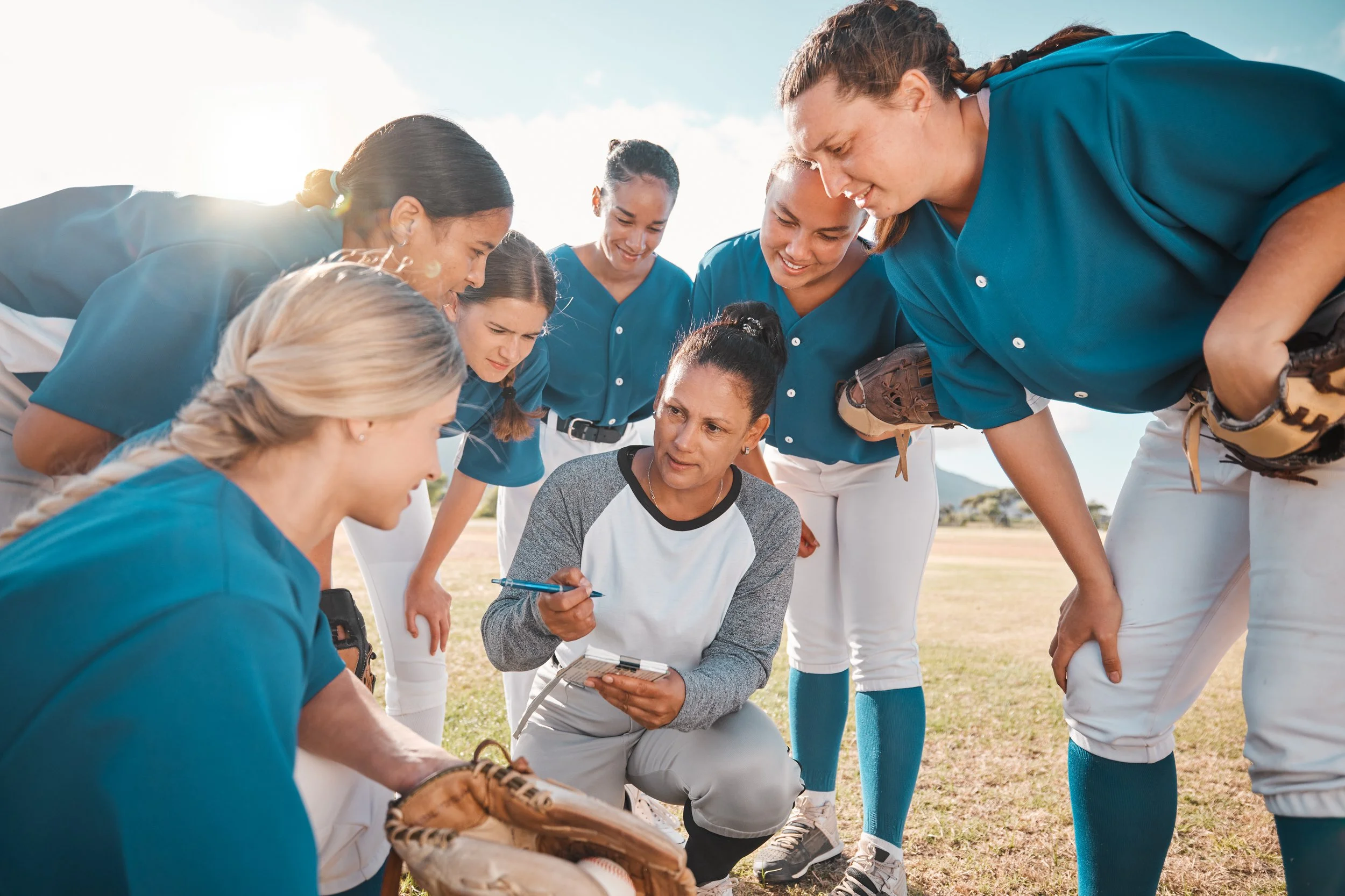 Softball coach instructs team during outdoor practice on grass field on sunny day.