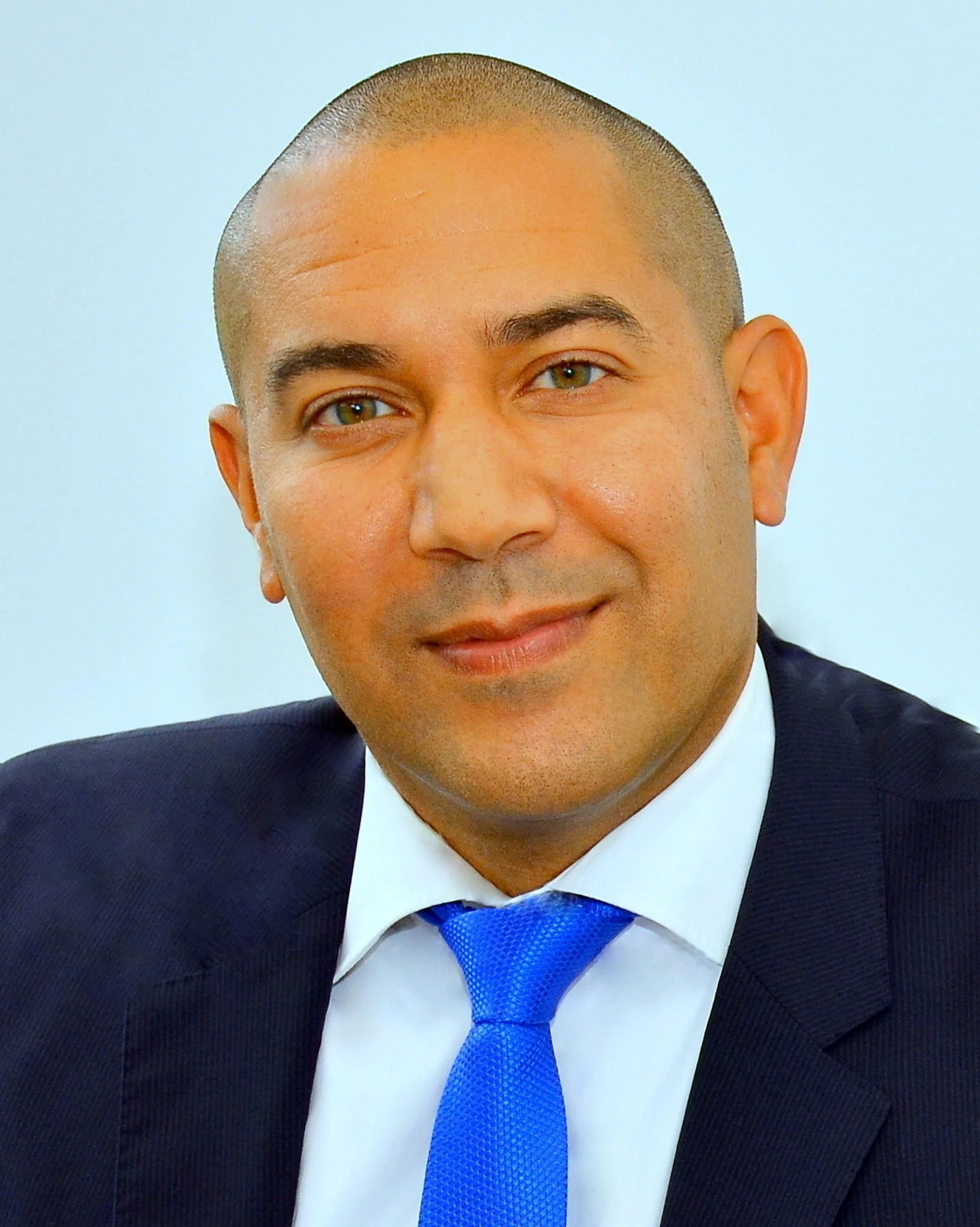 A man with a close-cropped haircut, wearing a dark suit, white shirt, and blue tie, smiling slightly. Only part of his face is shown, against a light background.