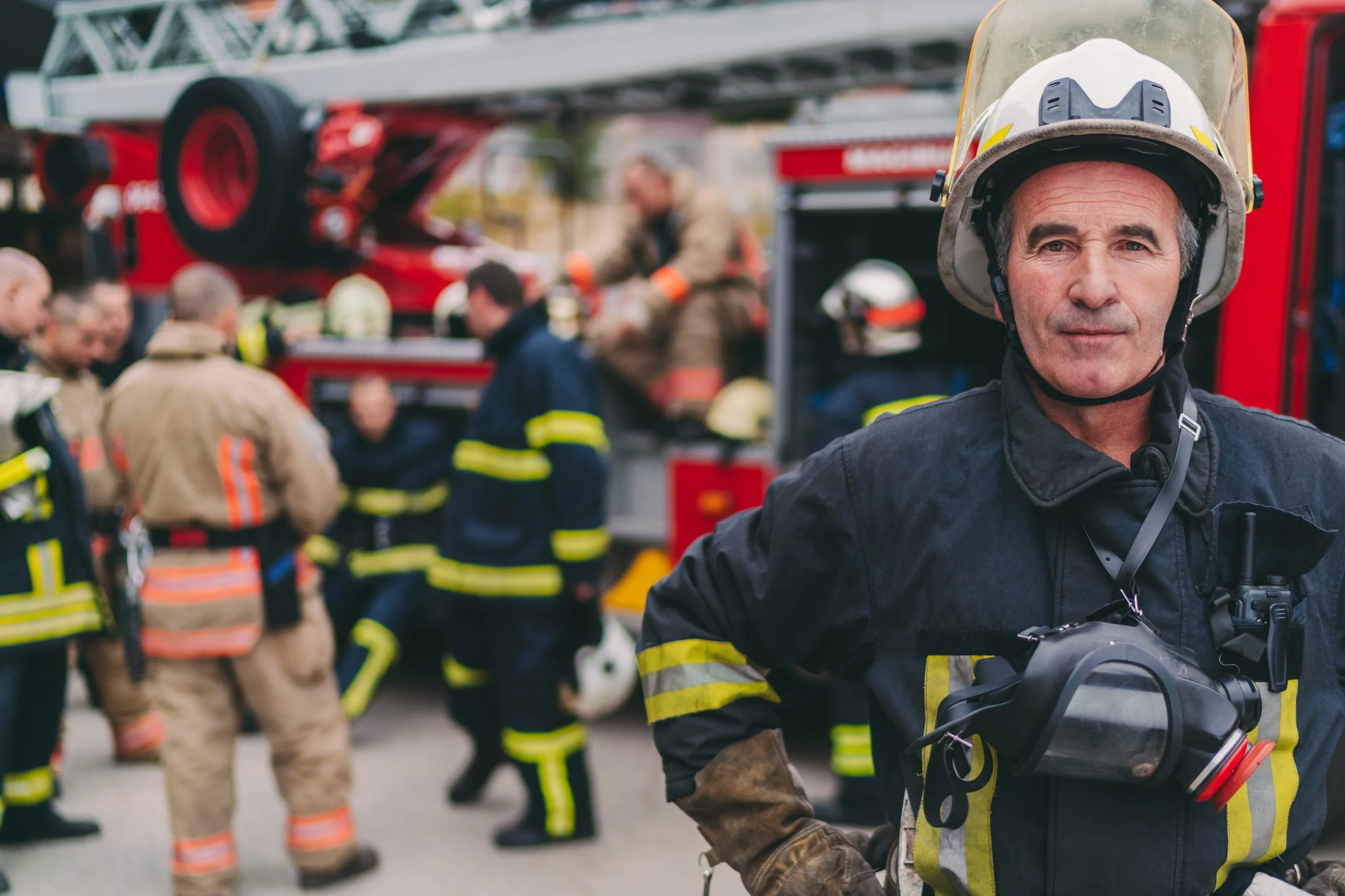 Firefighter wearing a helmet and protective gear standing in front of an emergency scene with other firefighters and a fire truck in the background.