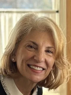 A smiling woman with shoulder-length blonde hair and light skin, indoors near a window with curtains.